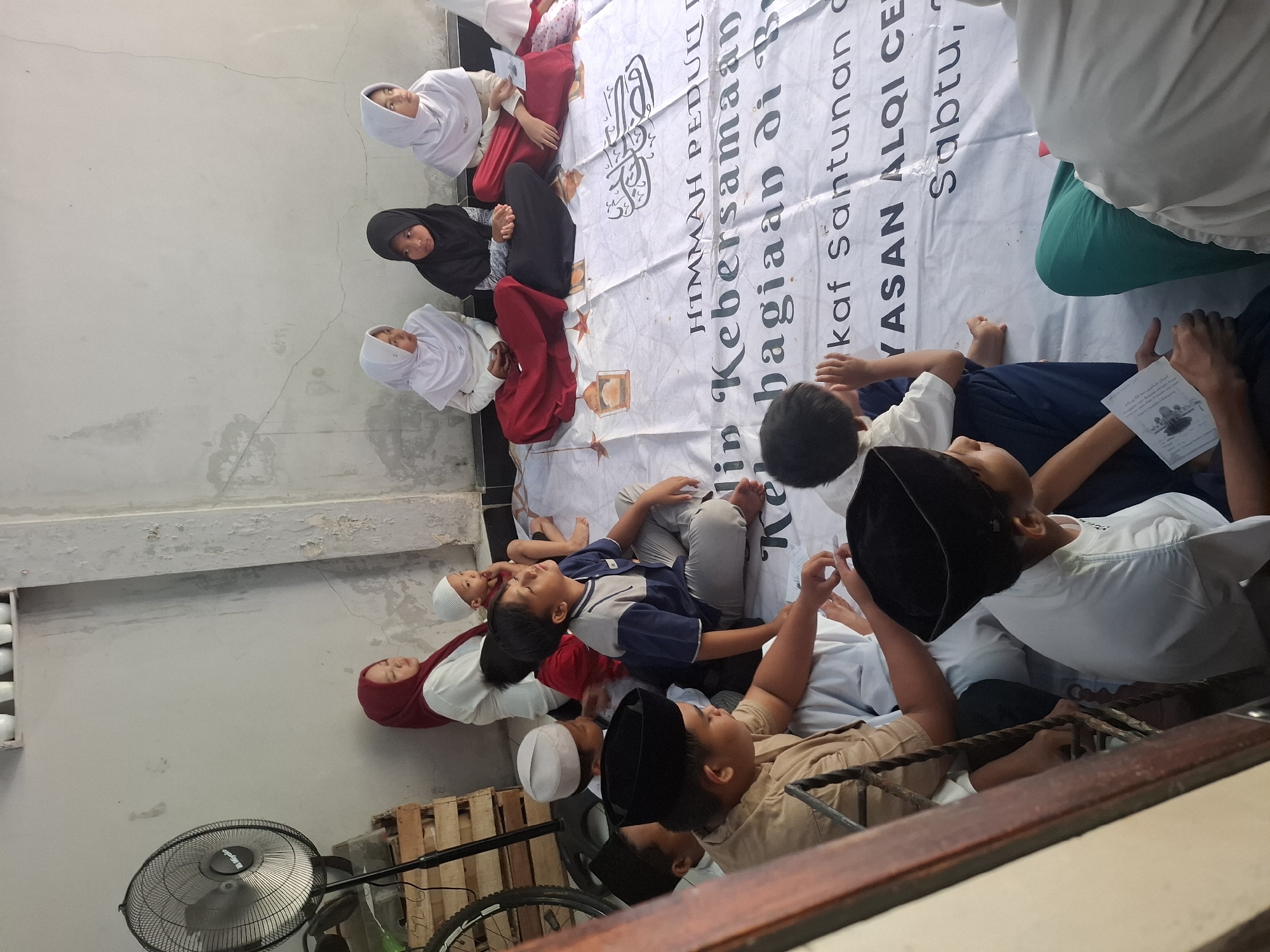 In the city of Babakan Madang, Indonesia, a group of children and a few adults are gathered on a white tarp spread on a tiled floor. The scene appears to be outdoors, with a pale, textured wall in the background. A tall, black electric fan is positioned to the left of the frame.
The group consists of approximately ten individuals, primarily children, dressed in modest attire, many wearing Islamic head coverings like hijabs and peci hats. Some of the children are seated cross-legged on the tarp, while others are in various stages of sitting or leaning.
A large white banner with text in both Arabic script and Latin characters is laid out on the ground, serving as the focal point for the gathering. The banner contains phrases such as "HIMMAH PEDULI" and "bersamaan di". There are also decorative elements on the banner resembling lanterns.
Several children are looking up or in different directions, while some appear to be engaged in conversation or listening to someone. One child in the foreground on the right is holding a small, folded paper or booklet. The overall atmosphere suggests a communal event, possibly a religious or charitable gathering, in an informal setting. The lighting indicates it is daytime, likely under natural light conditions.