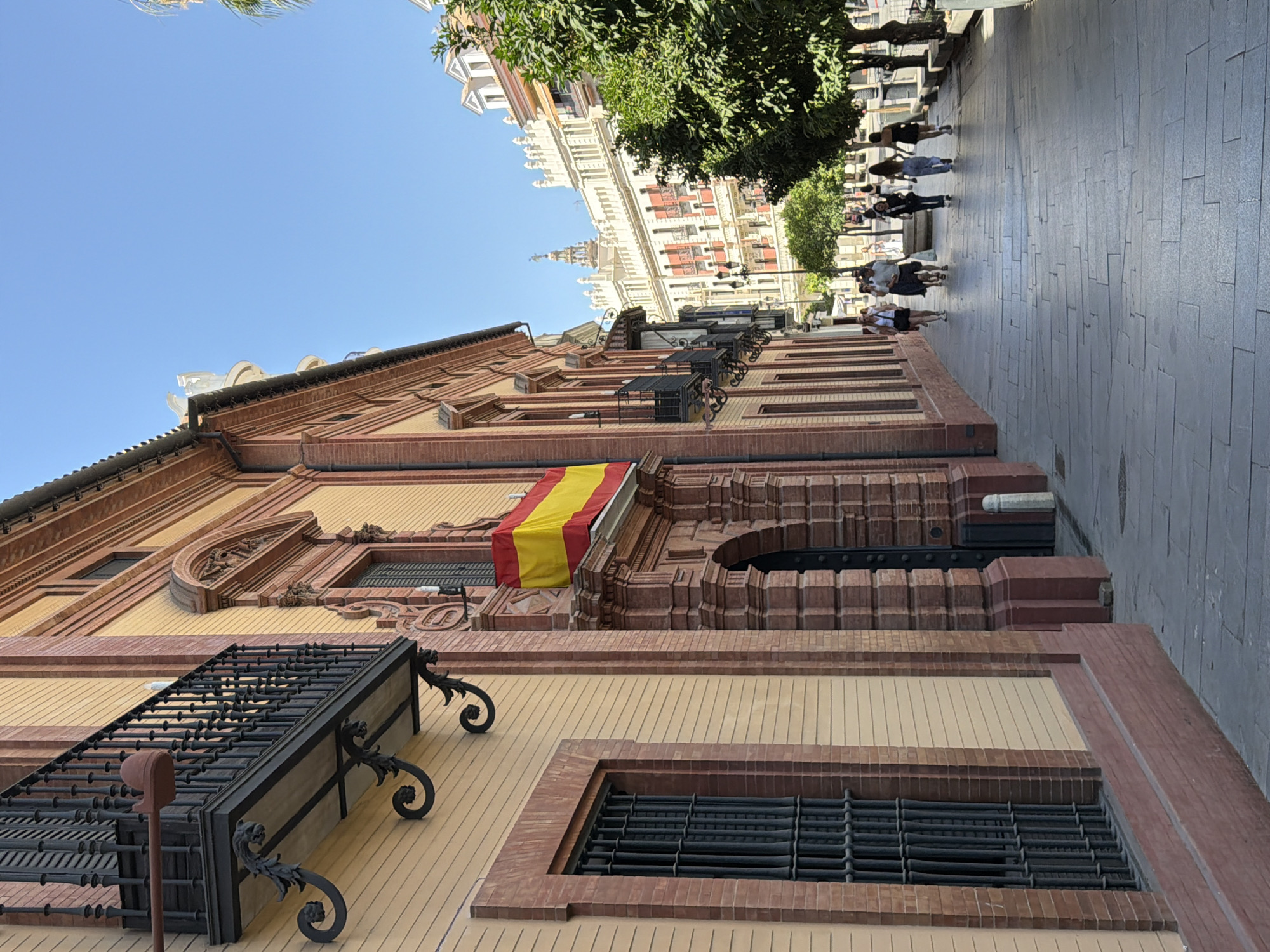 This is a photograph taken from a low angle, looking up at a building with ornate architectural details, likely a historical building in Seville, Spain. The building's facade is constructed with reddish-brown brick and features decorative elements in a lighter yellow material. There are several windows with black metal grates and intricate wrought-iron balconies with decorative scrolls. A Spanish flag, yellow and red, is draped over a part of the building.
In the background, a street lined with trees and other buildings is visible. The buildings in the distance appear to be tall and have classical architectural styles with white and red facades. A group of people are walking along the paved sidewalk on the right side of the image. The sky is clear and blue, indicating a sunny day. The time of day appears to be late morning or early afternoon, suggested by the bright sunlight and the shadows cast. The overall mood is serene and picturesque, showcasing a glimpse of urban life in a historic Spanish city. No visible text is present in the image.