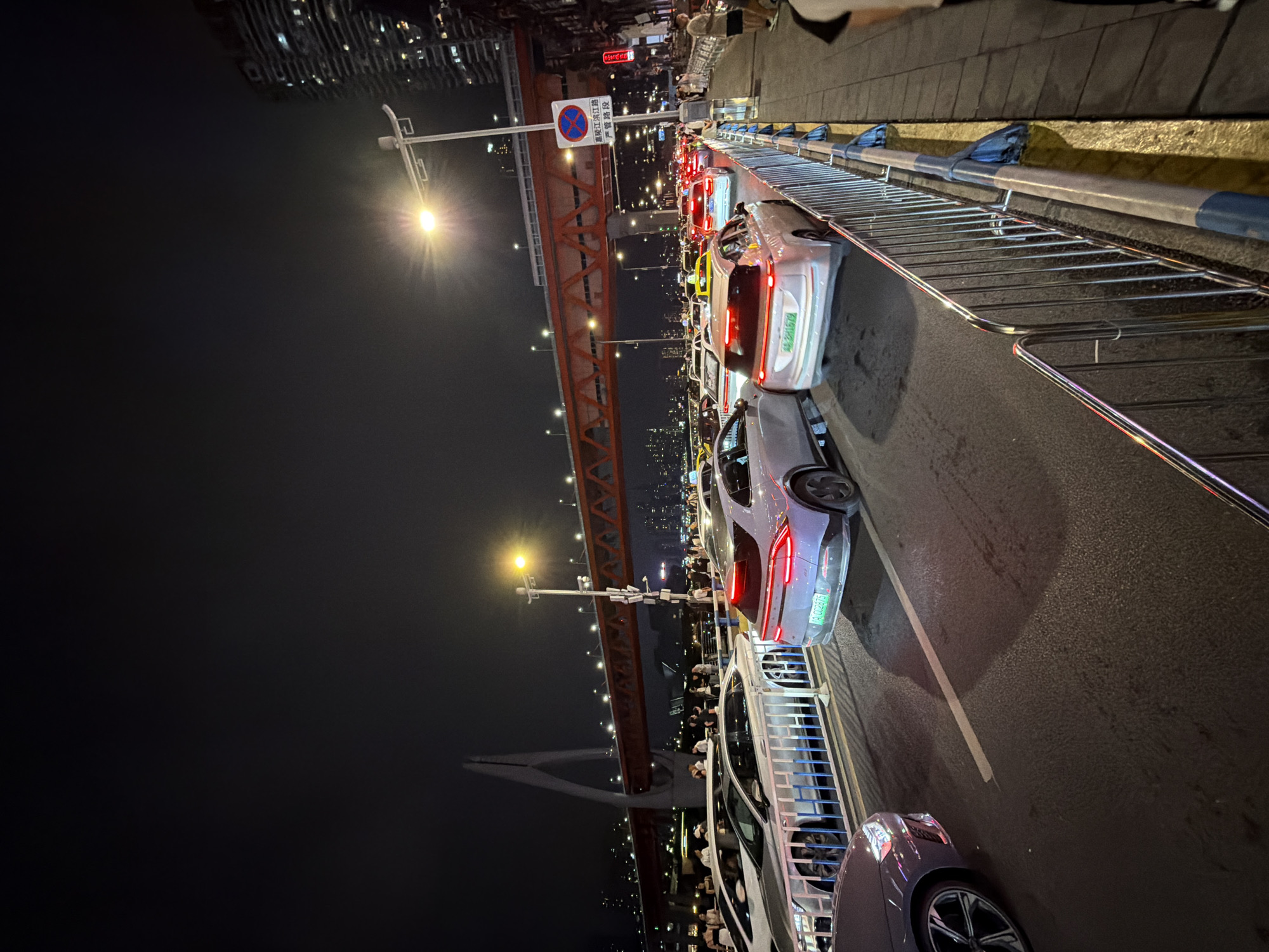 At night, on a bridge in Yuzhong District, China, traffic is backed up. Multiple lanes of cars, including sedans and SUVs, are stationary on the asphalt road. People are visible on a pedestrian walkway on the right side of the image. A large suspension bridge with red structural elements and illuminated lights spans the background, with city lights and buildings visible on the horizon. A street lamp illuminates a portion of the scene.