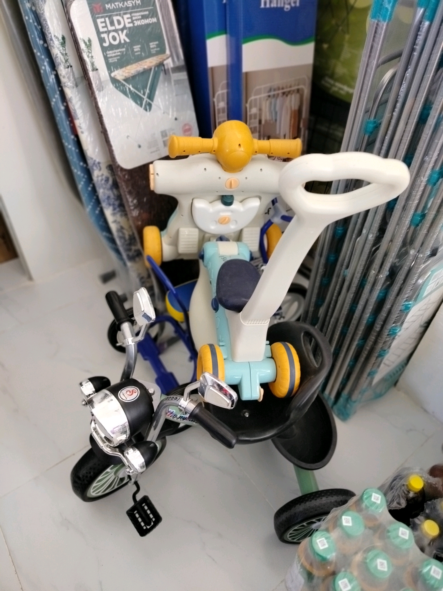 A close-up, eye-level shot showcases a predominantly white and blue children's tricycle with yellow accents, positioned on a light-colored tiled floor. The tricycle features a white handlebar with a large, white grip, a blue seat, and yellow wheels. In the background, several items are visible, including what appear to be folded clothes or fabrics in plastic packaging, as well as a tall, blue object with green accents. To the right, a stack of long, metallic rods, possibly part of a drying rack, is partially obscured by the tricycle. In the lower right corner, several plastic bottles with green caps and yellow lids, packaged together, are visible. The lighting is bright, casting minimal shadows. The image does not contain any individuals or any clear indication of the specific location within the city of Көлкент, Kazakhstan, beyond it being an indoor setting.