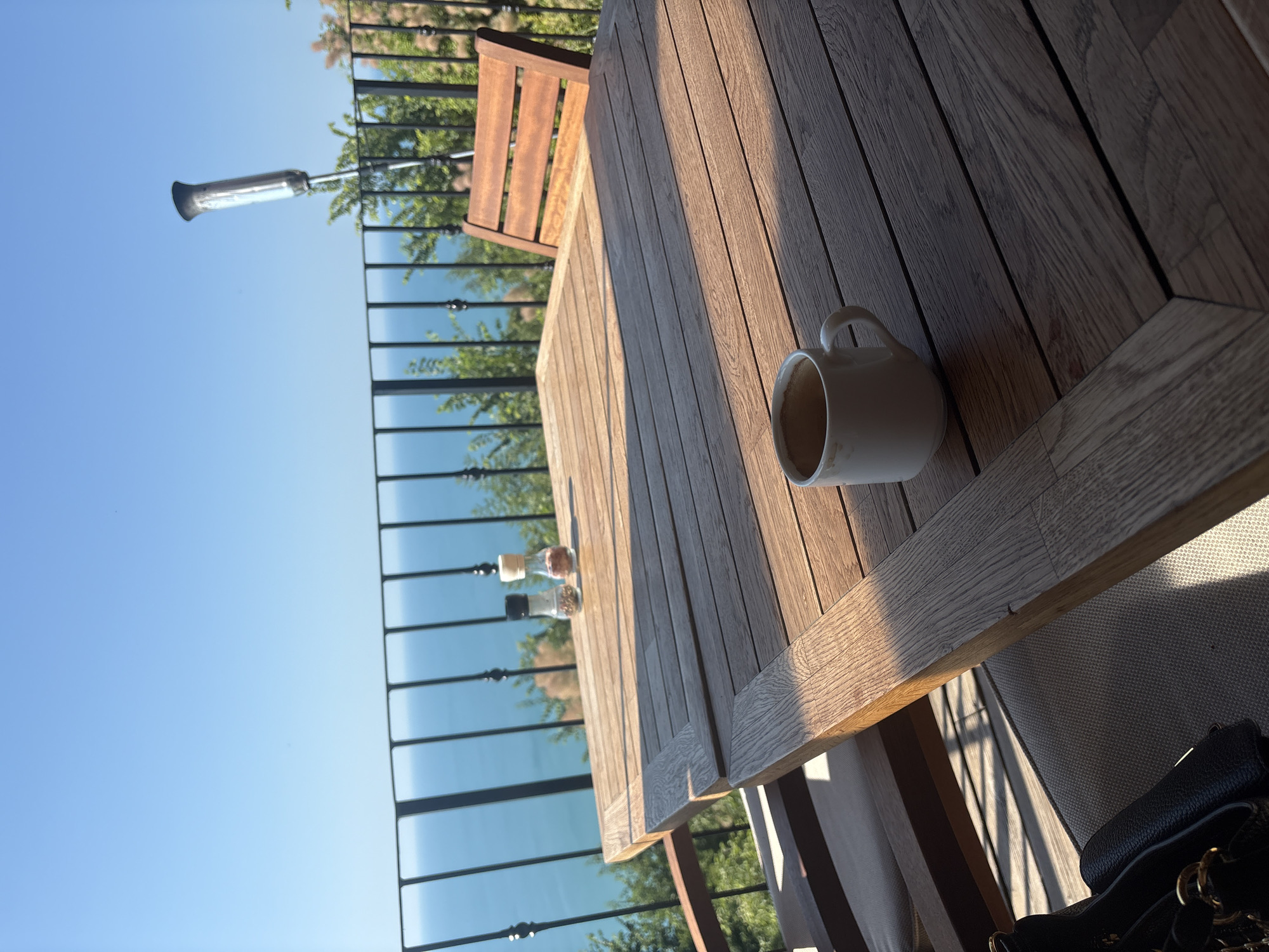 A close-up, eye-level shot captures a wooden table set against a backdrop of the ocean and a clear blue sky. On the table, a white mug with a light brown rim sits to the right, with a salt and pepper shaker set visible in the background. Two wooden chairs are partially visible, one to the right and another to the left, with upholstered seats. A black handbag rests on the left side of the frame. A black metal railing runs horizontally across the mid-ground, separating the table from the expansive ocean view. Lush green foliage is visible behind the railing, and a tall, metallic heater extends upwards into the sky on the far right. The lighting suggests it is daytime, with strong shadows cast across the table.
