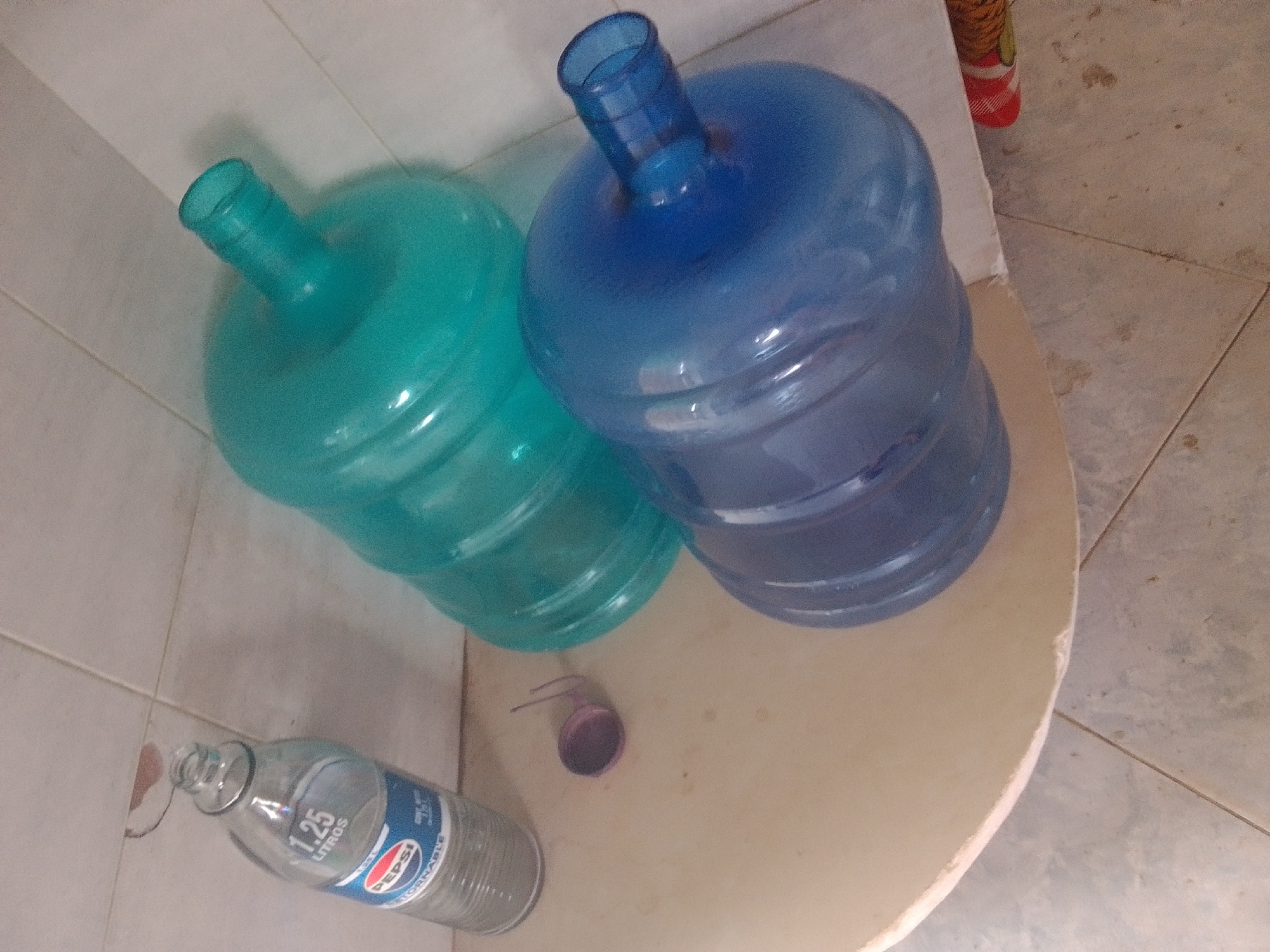 The image shows two large water cooler jugs, one turquoise and one blue, resting on a light-colored surface that appears to be a tabletop or counter. The jugs are placed side-by-side. In the foreground, a clear glass bottle with a blue and red Pepsi logo lies on its side. The bottle has a label that reads "1.25 LITROS" and "PEPSI". Next to the water jugs, there is a small, purple, cap-like object with a thin handle. The background consists of tiled walls and floor, suggesting an indoor setting, possibly a kitchen or utility area. The lighting suggests it's daytime. There are no people visible in the image.
