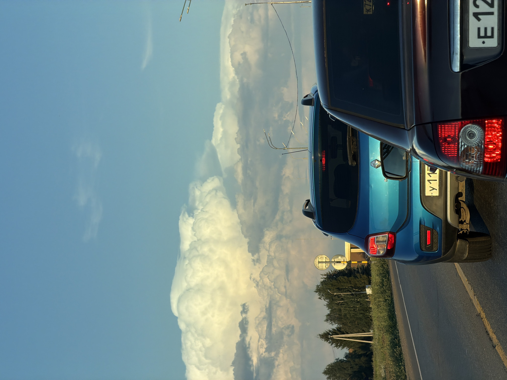 The image shows a traffic scene with two cars on a road. The sky is blue with large, dramatic clouds. The foreground car is dark brown and its rear is visible. Behind it is a bright blue car. The road appears to be asphalt with yellow lane markings. Trees and utility poles are visible on the left side of the road, along with traffic signs. The scene is likely in a semi-urban or rural area, possibly on the outskirts of Derzhavino, Russia, given the presence of the license plates. There are no people visible in the image.