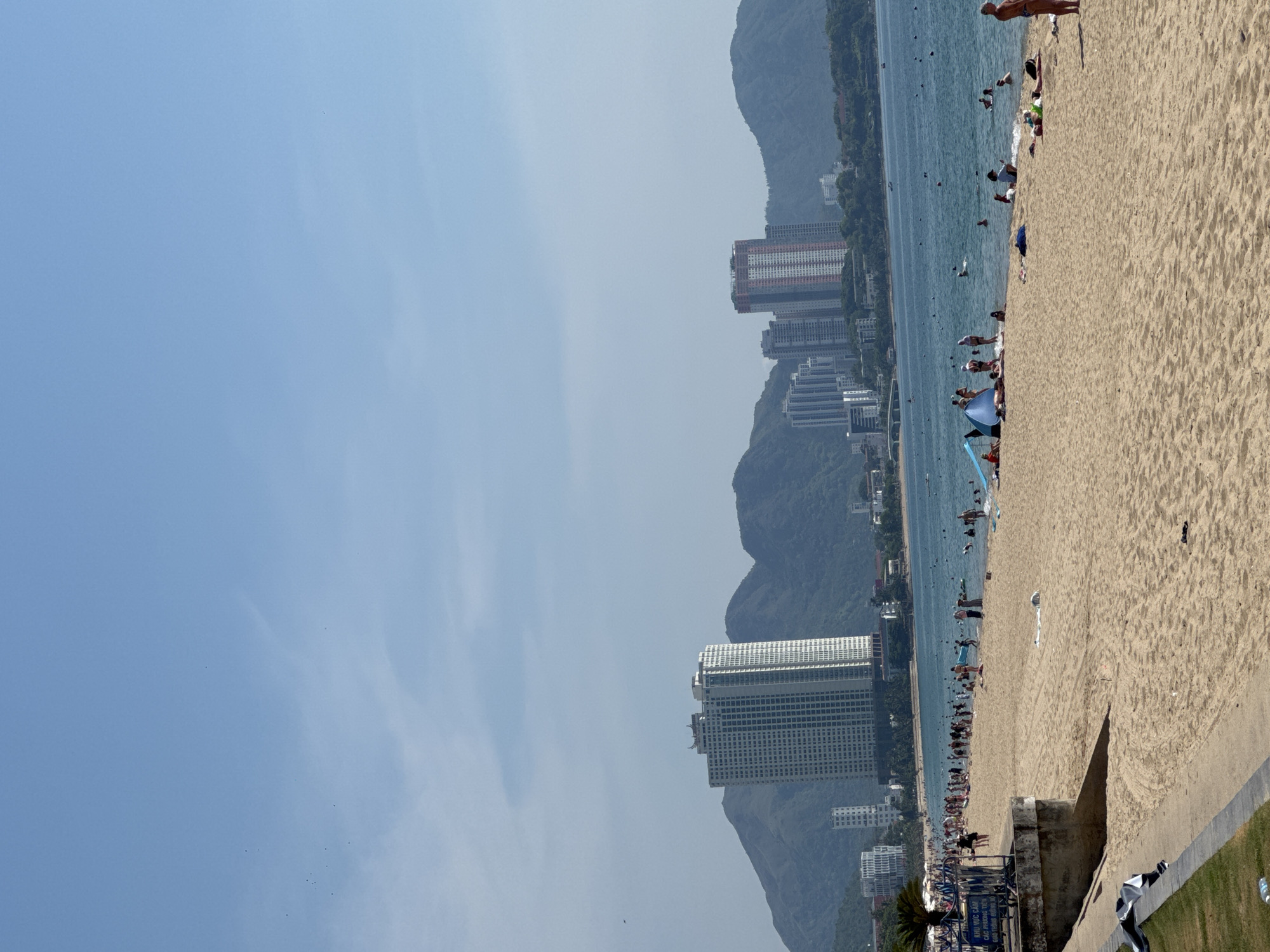 The image shows a crowded beach in Nha Trang, Vietnam, with numerous people sunbathing, swimming, and relaxing. The beach is wide and sandy, with the ocean in the background. Tall buildings and mountains can be seen in the distance, indicating an urban coastal environment. The sky is clear and blue. People are dressed in swimwear, and some are using umbrellas or beach tents for shade. The overall atmosphere appears to be one of leisure and recreation.