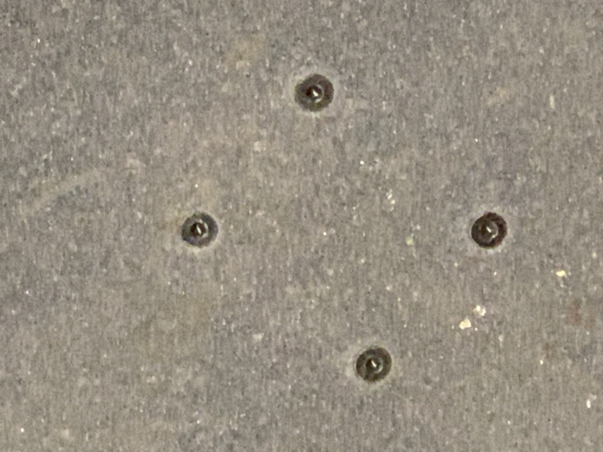 The image is a close-up, overhead shot of a textured, grey surface with five small, circular objects embedded in it. These objects appear to be screws or rivets, each with a dark, recessed head and a visible slot or cross-shaped indentation. They are arranged in a rough cross or plus shape, with one centered at the top, one at the bottom, one to the left, one to the right, and one slightly off to the bottom left. The surface itself has a speckled appearance, with lighter and darker grey tones, and some small, reflective white specks are visible, possibly dust or small particles. The lighting is even, and there are no shadows cast by the objects. The image does not provide any context regarding its specific location, and therefore, its placement in Barcelona, Spain, cannot be determined from the visual information alone.
