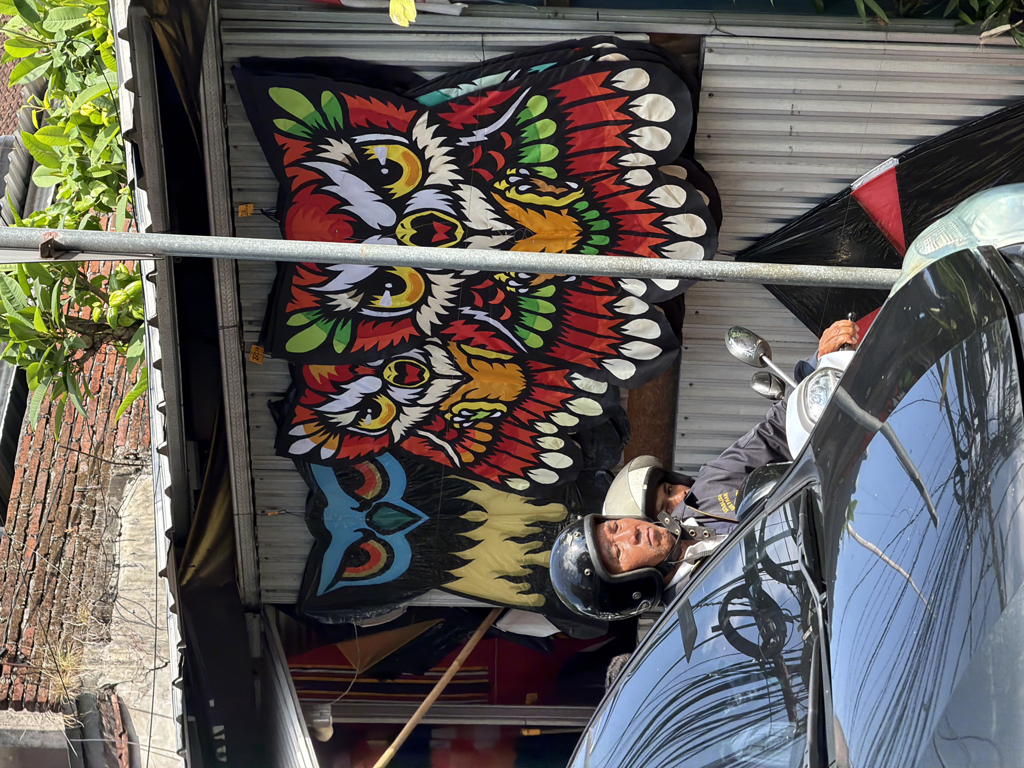 In Ubud District, Indonesia, a man wearing a black helmet is riding a scooter, with another person behind him also wearing a helmet. They are in front of a structure with a corrugated metal wall where several large, colorful kites shaped like owls are displayed. The kites feature intricate designs with prominent eyes, beaks, and feather patterns in red, yellow, green, and white against a black background. A dark-colored car is parked in front of the scooter, its hood reflecting the surroundings. The environment appears to be an outdoor market or shop, with overhead awnings and greenery visible. The overall scene captures a street-level view in what seems to be a commercial area of Ubud.