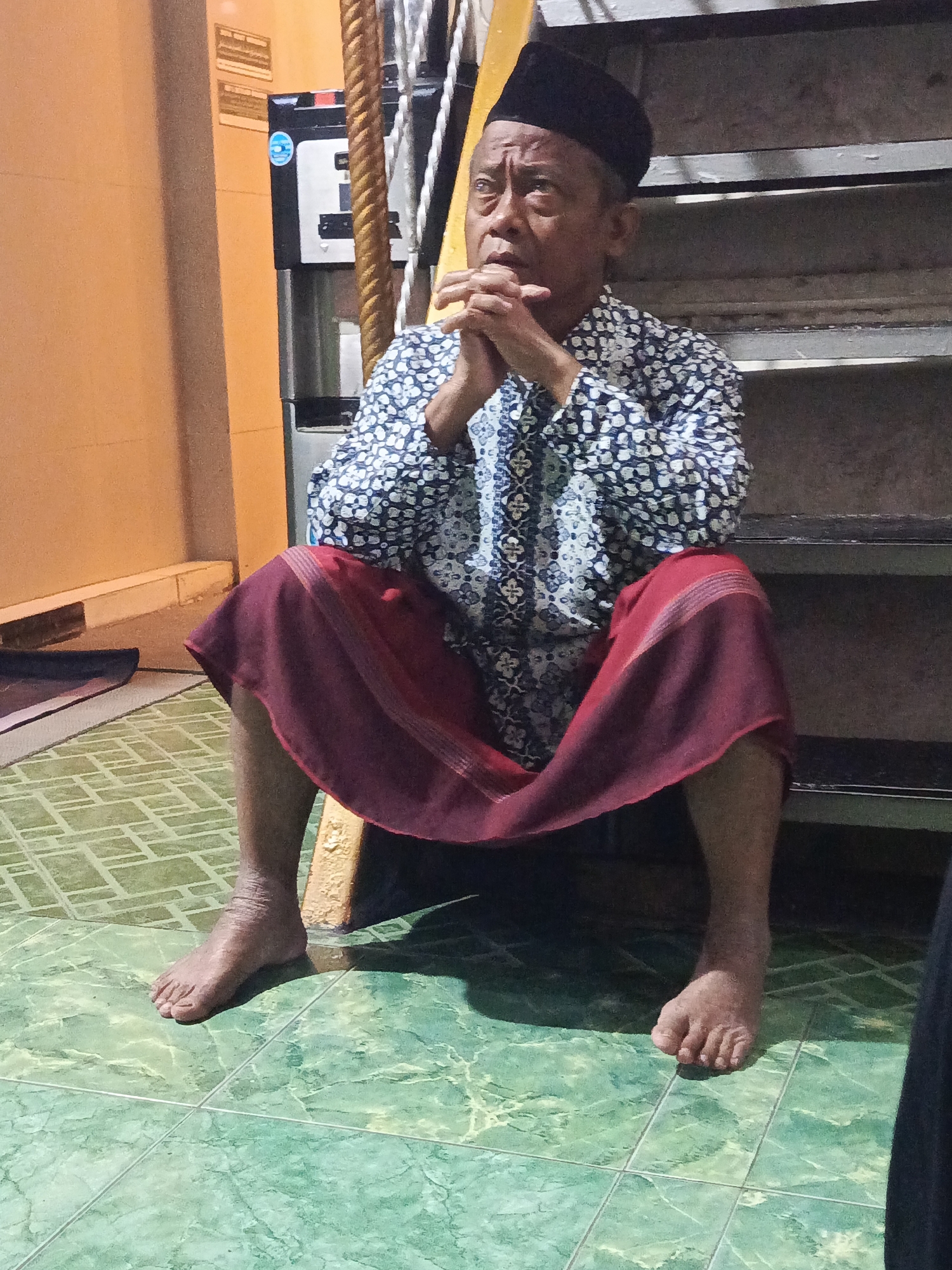 An older man wearing a black peci and a patterned blue and white shirt is seated on a step. He has a maroon sarong wrapped around his lower body. His hands are clasped together in front of him, and he is looking upwards with a pensive expression. The man is sitting on a concrete step, with other steps visible behind him. The floor is covered in green tiles. To the left of the frame, there is an orange wall with a water dispenser against it. The image appears to have been taken indoors.