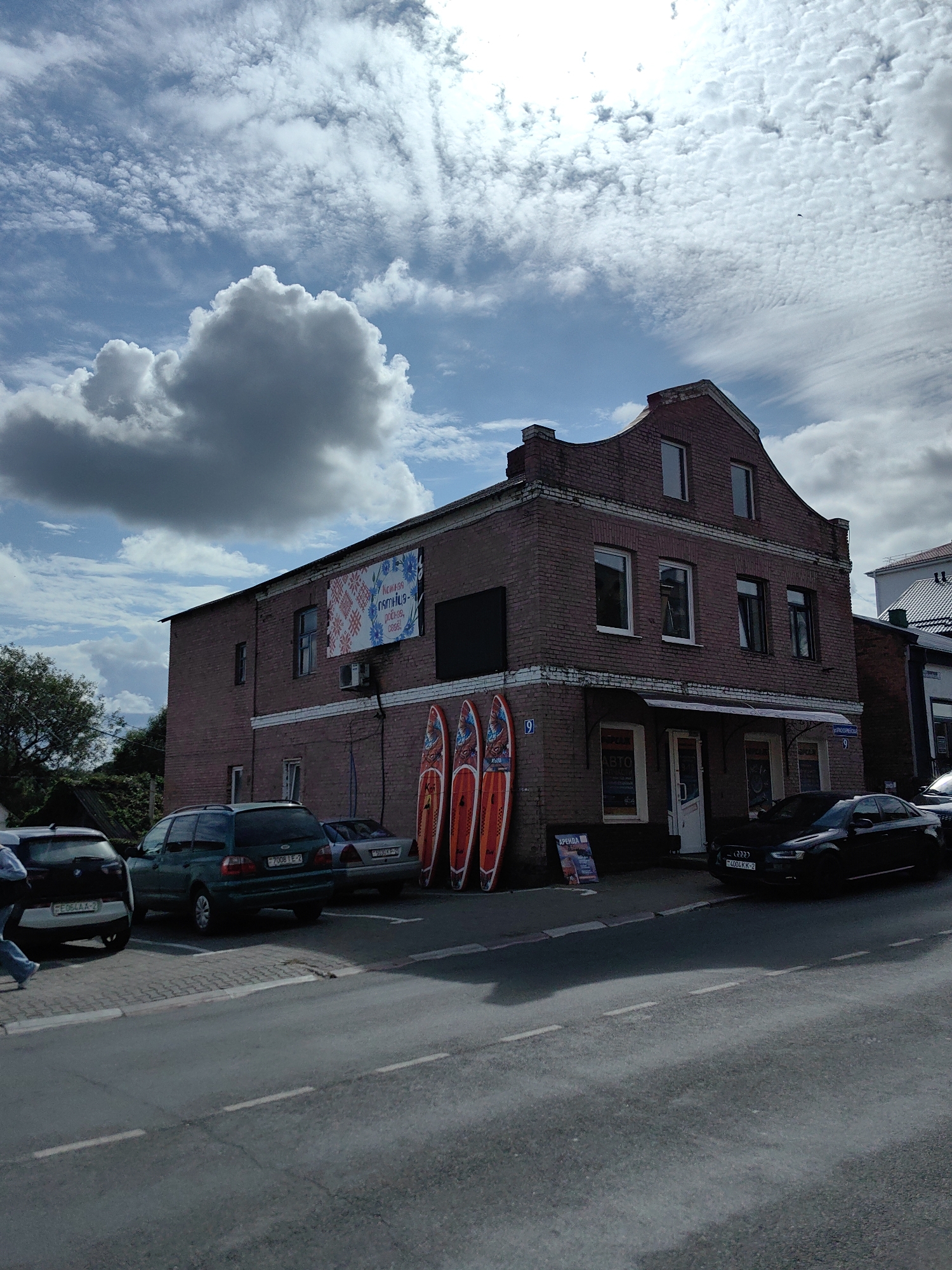 The image depicts a brick building in the city of Pastavy, Belarus, with a prominent sign on its facade. The building appears to be a commercial establishment, with several windows and an awning over the entrance. In front of the building, there are three stand-up paddleboards leaning against the wall, suggesting activities related to water sports or rentals. Several cars are parked along the street in front of the building. The sky is partly cloudy, with a mix of sunlight and shadow casting across the scene. The overall atmosphere suggests a typical day in a town, with commercial activity and outdoor elements.