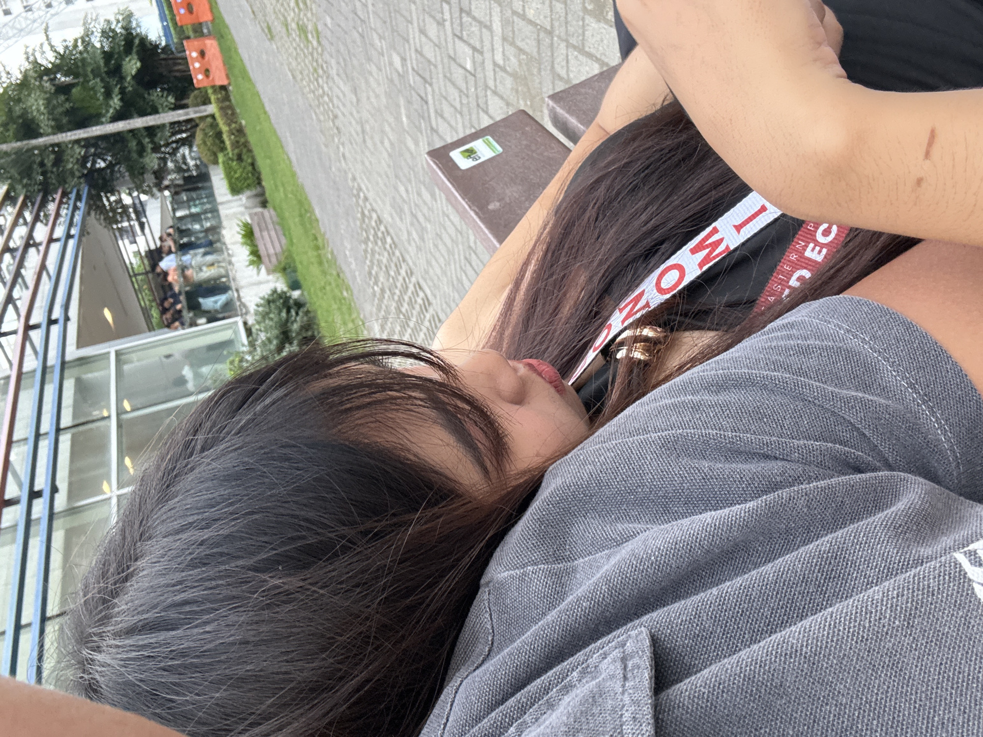 A young Asian woman is resting her head on a person's shoulder. The woman has dark hair and is wearing a gray shirt. She also has a headband on with the words "EASTERN LEADERSHIP DEC". The person she is leaning on has their arm around her. The location appears to be outdoors, possibly in Davao City, Philippines, based on the architecture and foliage visible in the background. There are buildings with large windows, trees, and possibly street furniture in the background. The scene suggests a moment of relaxation or intimacy between the two individuals.