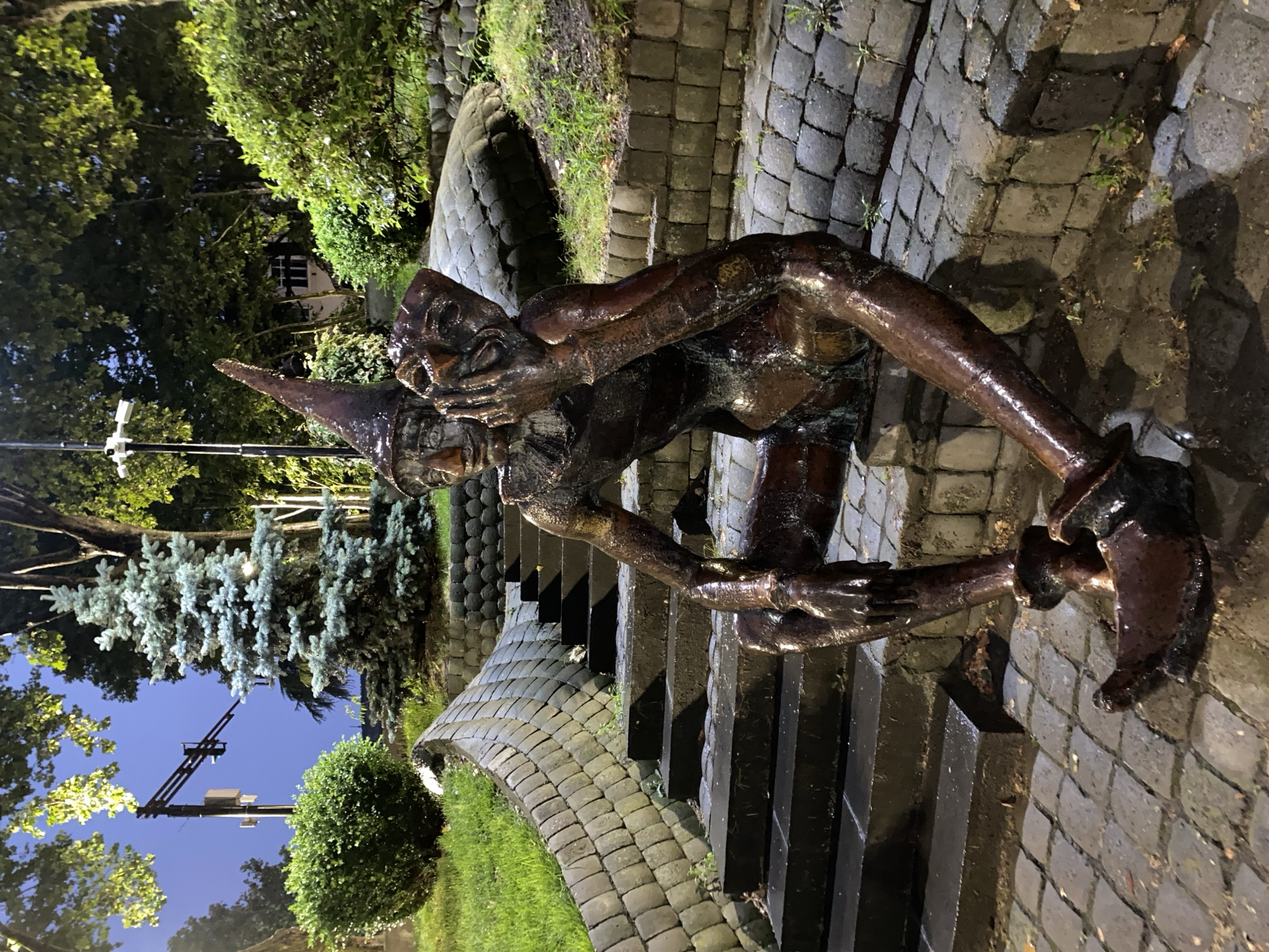 The image depicts a bronze sculpture of a mythical creature, possibly a gnome or a goblin, kneeling on a stone pathway at night. The creature has a pointed hat, a long nose, and appears to be holding its head in its hands, with a distressed or pained expression. Its body is hunched over, and its limbs are twisted in an awkward pose. The sculpture is dark and appears wet, suggesting recent rain or a damp environment.
The setting is an outdoor area with cobblestone paving and a curved retaining wall made of similar stones. Grass and lush green bushes surround the pathway and wall, with a few evergreen trees visible in the background, one of which is a light blue spruce. The lighting suggests it is nighttime, with artificial lights illuminating the sculpture and the surrounding area. A streetlamp with a green light is visible in the distance, along with what appears to be power lines. The sky is dark blue, indicating it's either late evening or early morning.
The overall mood of the image is somewhat somber and mysterious, enhanced by the nighttime setting and the creature's apparent distress. The wet surfaces of the cobblestones and sculpture add a reflective quality, emphasizing the low light conditions. No text is visible in the image. The location context is Tbilisi, Georgia.