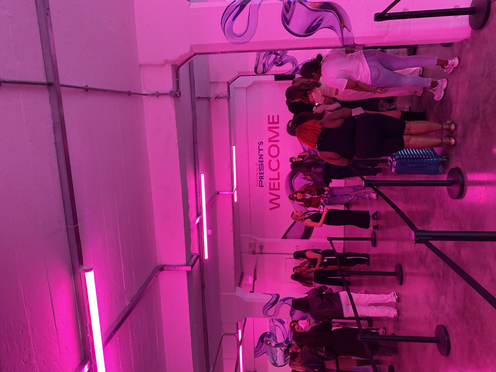 This media file captures an indoor scene, likely a waiting area or entrance to an event, illuminated by prominent, vibrant pink fluorescent strip lights that cast a strong magenta hue across the entire space. The floor appears to be a grey, possibly concrete, surface.
A queue of approximately a dozen people, predominantly women of various ages and ethnicities, are standing in line, marked by black retractable belt barriers (stanchions). They are casually dressed, with some individuals looking at their phones, while others are engaging in conversation or patiently waiting. One person has a blue hard-shell suitcase at their feet. The overall mood appears neutral and anticipatory.
Architectural details include exposed silver-colored conduits running along the walls and ceiling. The walls themselves are light-colored, tinted pink by the lights, and feature abstract, wavy, reflective purple decorations.
Prominently displayed on a wall, oriented vertically, is visible text: "Supergang ^ PRESENTS" followed by "WELCOME". This signage, along with the queue, strongly suggests an organized event or attraction. The scene is artificially lit, making the time of day indeterminate. Based on the provided context, this event is located in London, United Kingdom.