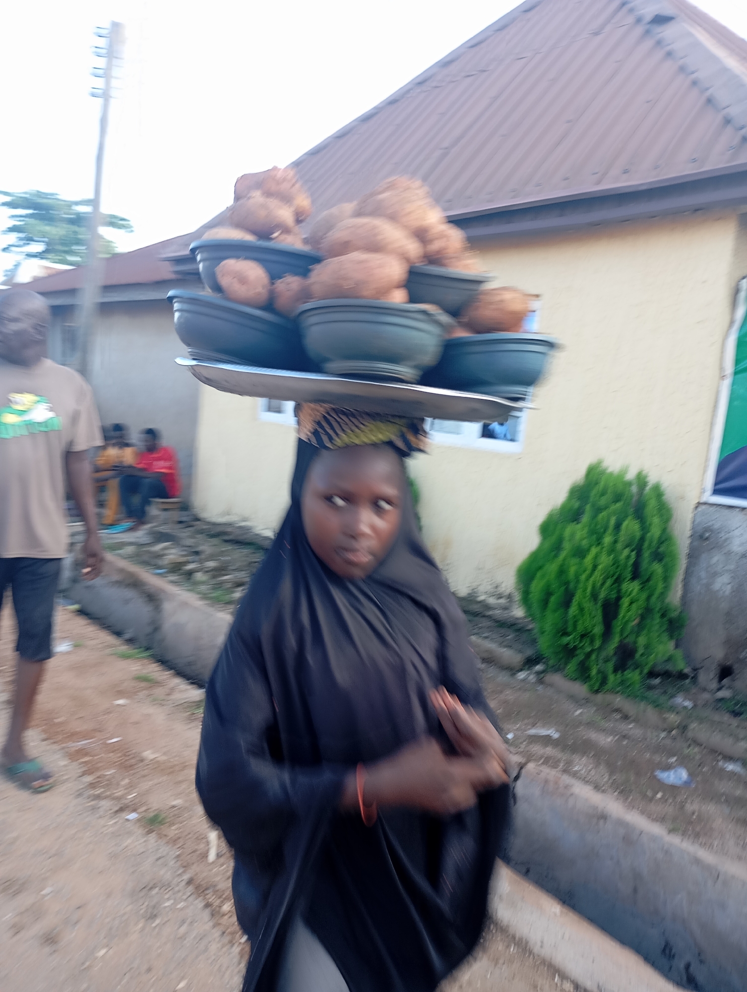 In Shendam, Nigeria, a young girl wearing a black hijab balances a tray of yams on her head. She is dressed in a black abaya and is walking, with her hands clasped in front of her. Behind her, a man in a tan t-shirt and shorts walks away from the camera, his left arm down and his right hand slightly raised. To the left of the girl, two people are seated, one in an orange shirt and the other in a red shirt. A green bush is visible to the right of the girl. The ground is dirt, and a concrete drainage channel runs along the left side of the image. A building with a brown roof and light yellow walls is visible in the background. A utility pole stands to the left of the frame.
