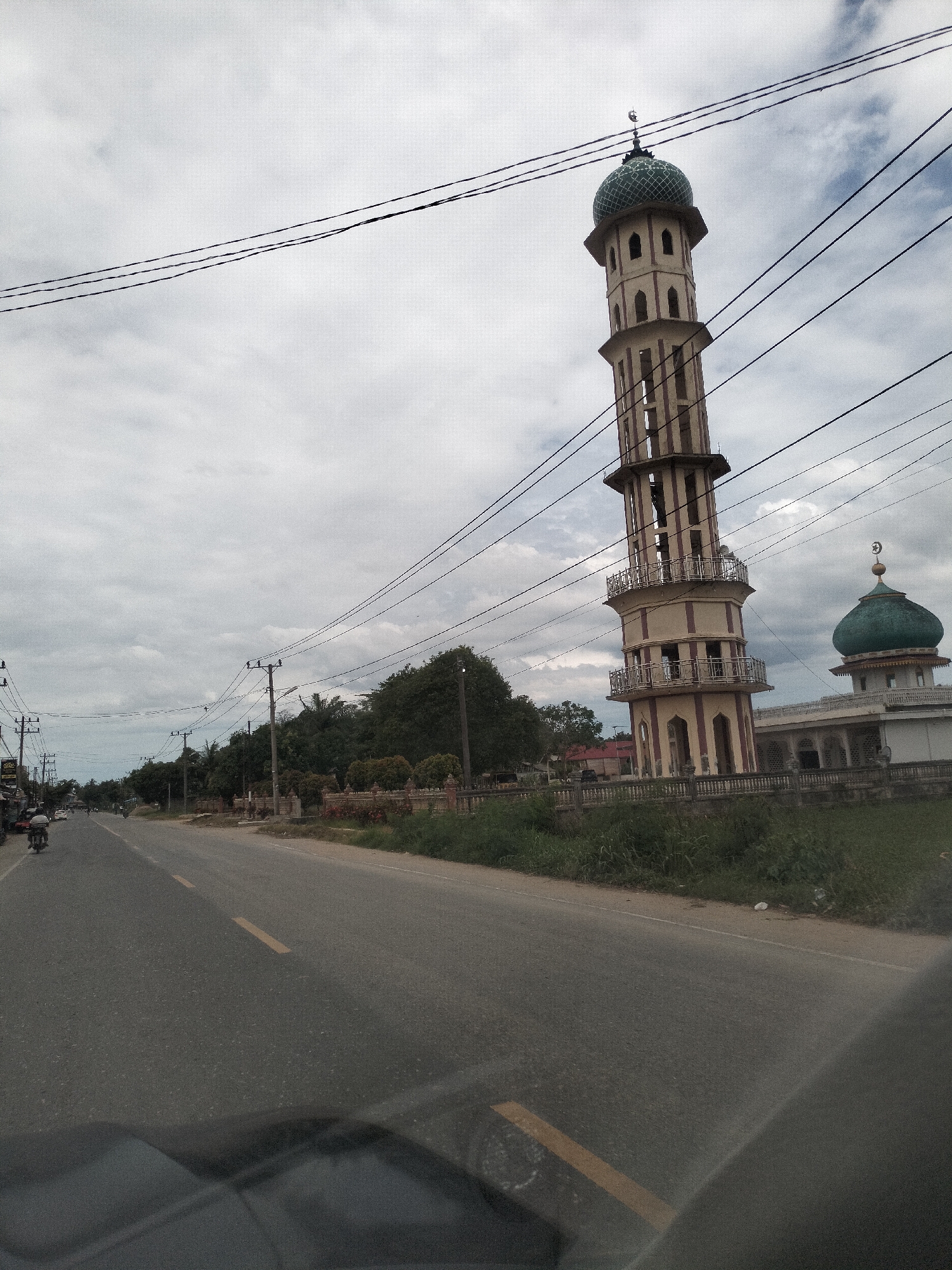 The image captures a street scene in Putoh Sa, Indonesia, under an overcast sky. A tall, multi-tiered minaret with a green dome stands prominently on the right side of the road, adjacent to a mosque with a matching green dome. Several power lines stretch across the frame, supported by utility poles lining the road. The asphalt road is marked with a yellow line and appears to be a two-lane street. On the left side of the road, there are some buildings, a few motorcycles, and a person on a motorcycle. Lush green vegetation, including trees and bushes, borders the road and the mosque complex. The foreground shows a reflection of the interior of a vehicle, suggesting the image was taken from a car.