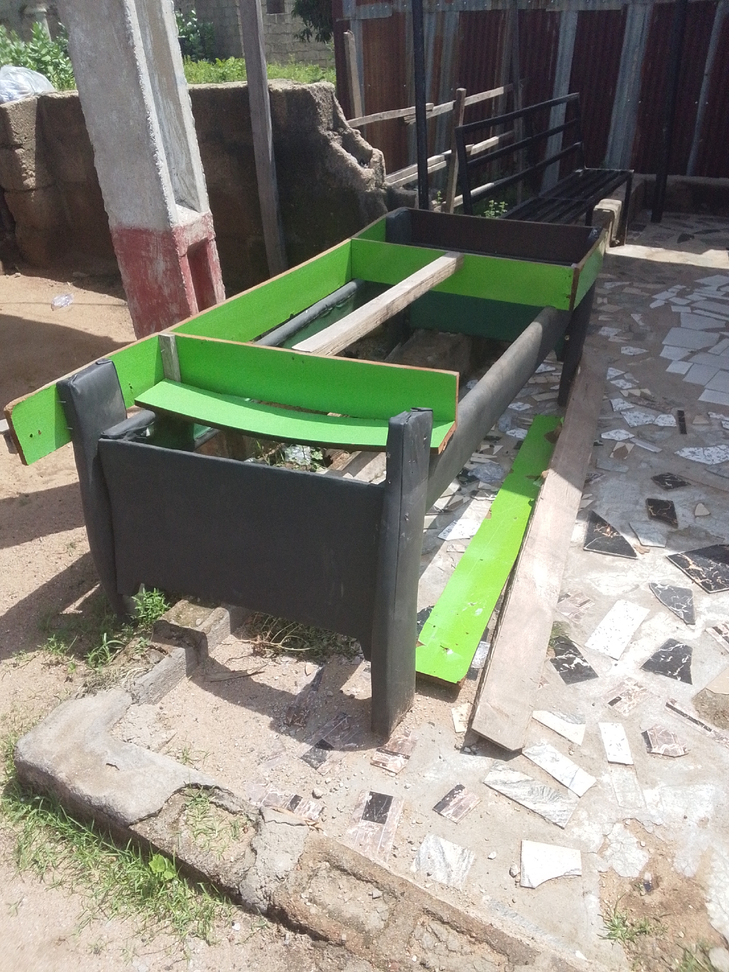 The image shows an outdoor scene in Jalingo, Nigeria. In the foreground is a partially constructed wooden structure, likely a bed frame, painted black and vibrant green. The frame appears to be a DIY project, with visible screws and raw wood. The surrounding area is a rough, uneven ground covered with dirt, scattered concrete debris, and some small patches of grass. Some mosaic tiles are also present on the ground, suggesting a paved area nearby that has been disturbed.
In the background, a weathered, corrugated metal fence runs horizontally across the frame, suggesting a boundary or enclosure. Behind the fence, some greenery and parts of buildings are visible, indicating a residential or semi-urban environment. A large, concrete pillar, painted red and white at its base, stands to the left of the frame, adding to the utilitarian and somewhat unfinished feel of the location. The lighting suggests it is daytime, with the sun casting shadows. There is no visible text in the image. The scene conveys a sense of ongoing construction or a work in progress in a warm, possibly dusty environment.