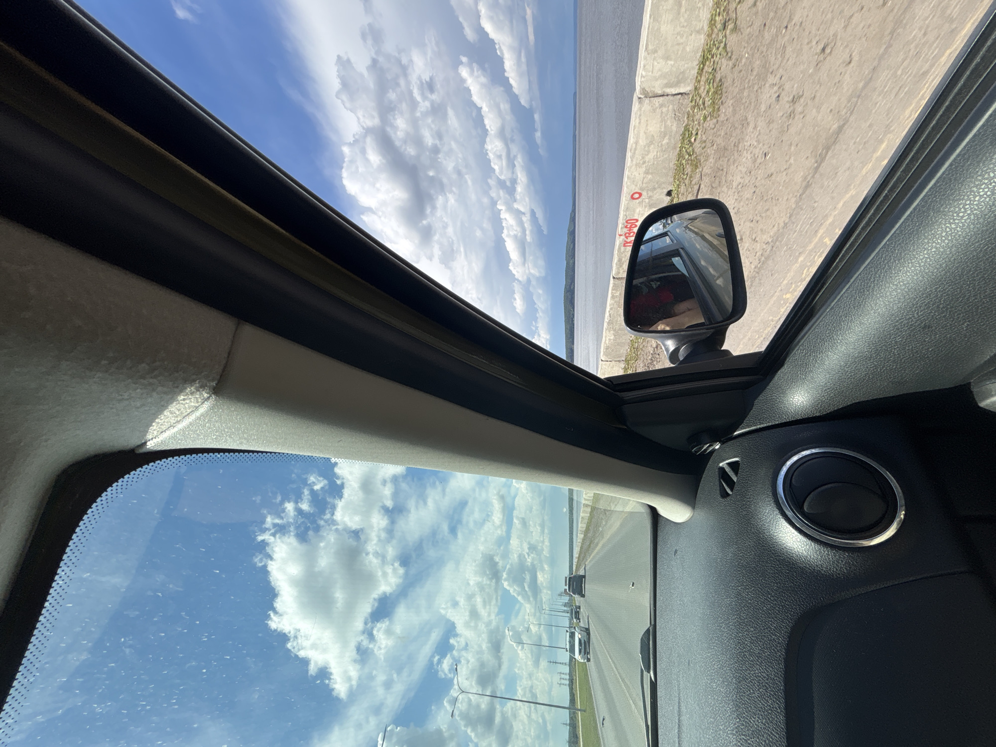 The image depicts a view from inside a car looking out through the windshield and an open sunroof. The scene is a daytime shot, with a bright blue sky filled with fluffy white clouds. The car is on a highway, indicated by the road stretching into the distance and streetlights lining the side. Several other cars are visible on the road ahead.
To the right of the car, a large body of water, likely a river or lake, is visible. A concrete embankment runs along the edge of the water, and some red markings with Cyrillic text are present on it, including what appears to be "ТК 13+60". A side-view mirror of the car is also visible, reflecting the interior of the car and a person with red clothing.
The interior of the car shows part of the dashboard with a ventilation grate and the fabric lining of the sunroof and windshield frame. The overall impression is a scenic drive on a clear day, possibly in or near Chaikovsky, Russia, given the location context provided. There are no strong emotional cues visible, beyond the general sense of a pleasant journey.