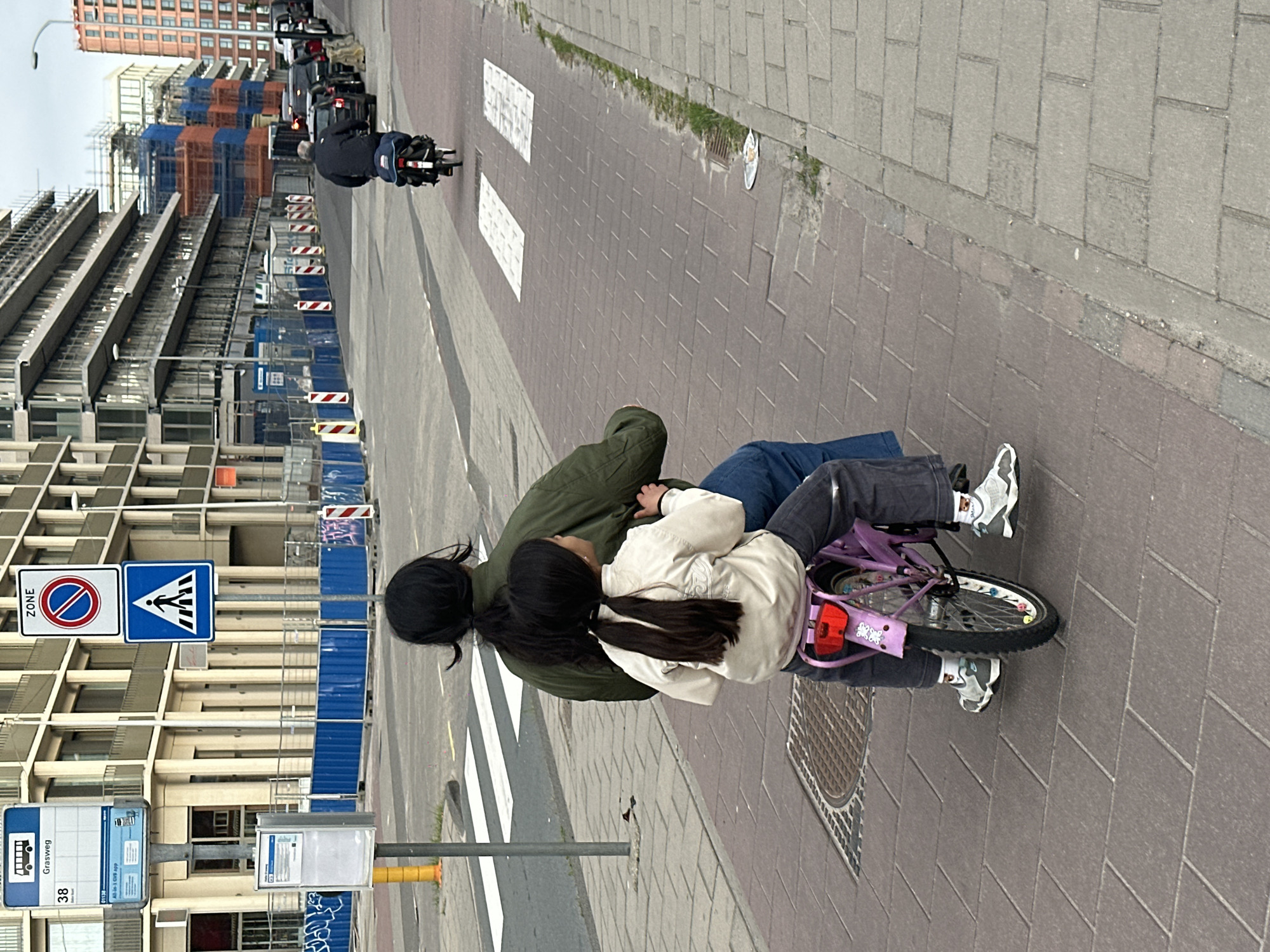 Two people are on a children’s bicycle on a street in Amsterdam, Netherlands. One person, wearing an olive-green jacket and blue jeans, is standing behind the bike, guiding it. The other person, wearing a cream-colored jacket and gray jeans, is seated on the bicycle. The bicycle is purple and pink with flower designs.
Behind them is a crosswalk with white stripes. Street signs, a bus stop sign, and a yellow pole are visible to the left. Construction is occurring on a building in the background, with blue metal fencing and orange netting. Additional buildings and vehicles are visible in the distance. The weather appears overcast. The ground is paved with rectangular gray bricks.