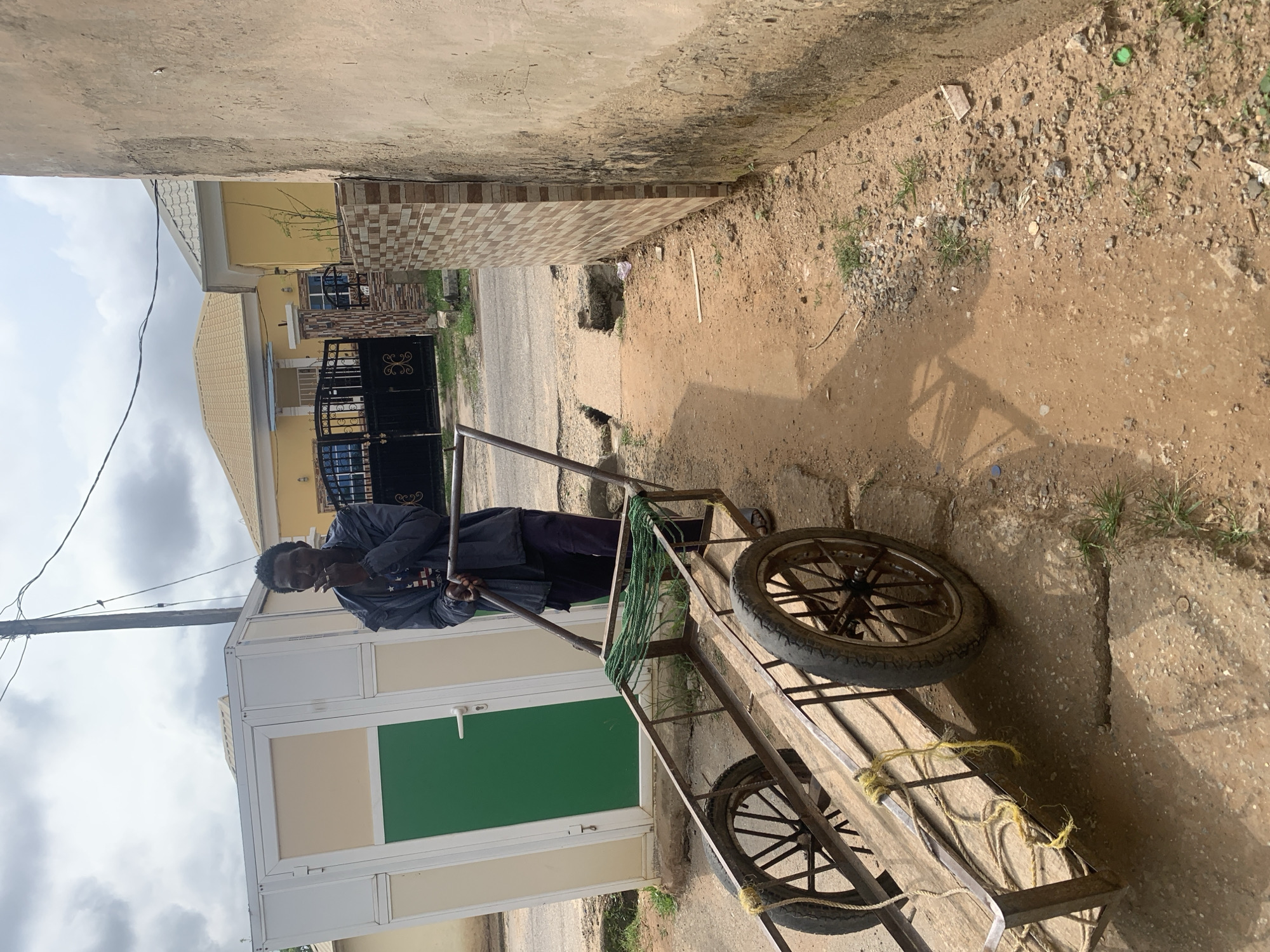 A man stands next to a metal and wooden handcart with two large spoked wheels in Abuja, Nigeria. The man is wearing a dark jacket over a t-shirt and dark pants. He is holding the handle of the cart and appears to be smoking. The handcart has a green net stretched across the front of its frame. Behind the man and cart is a modular building with a green door and beige panels. Further in the background, there are yellow houses with tiled roofs and a black metal gate. The ground is dirt and gravel. The scene is outdoors under a cloudy sky.