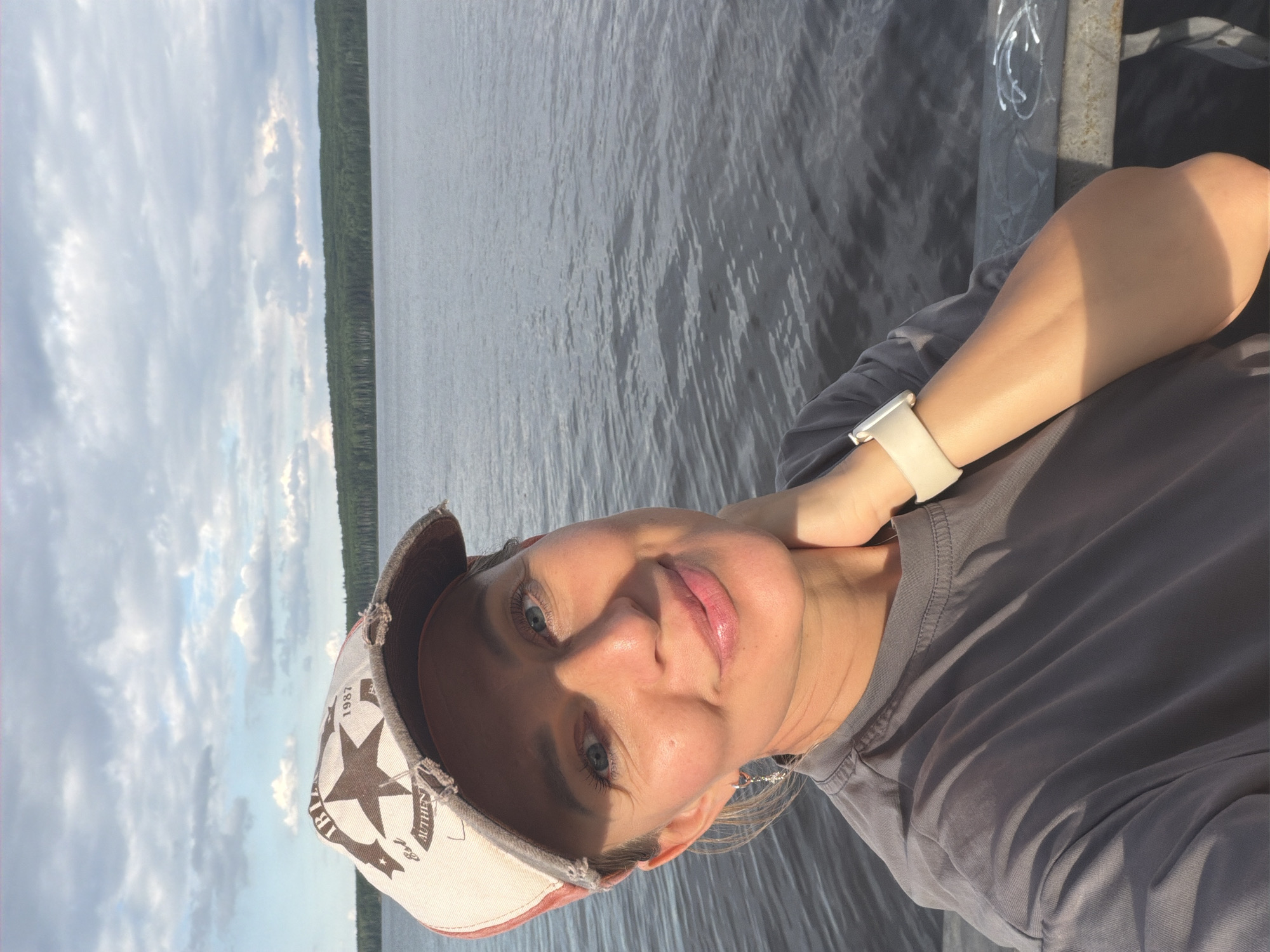 This is a portrait-oriented selfie of a woman wearing a grey t-shirt and a distressed baseball cap. The cap is light-colored with a brown brim and features a star graphic with text, including "1987" and "AUTHENTIC." The woman has light skin and blue eyes, and she is looking towards the camera with a slight smile. Her right arm is bent, and her hand is resting on her neck. She is wearing a light-colored smartwatch on her left wrist.
The background shows a large body of water with gentle ripples, suggesting a calm day. To the left of the water, a dense line of green trees is visible along the horizon, indicating a natural, possibly forested, landscape. The sky above is partly cloudy with dramatic, soft clouds and a mix of light and shadow, hinting at either early morning or late afternoon. The sunlight is casting distinct shadows on the woman's face and arm.
The overall mood of the image is relaxed and casual, with the subject appearing happy and comfortable. The scene suggests an outdoor setting, likely near a lake or river, possibly on a pier or a boat, given the presence of a metal railing visible on the right side of the frame, which has some graffiti on it. The location is identified as Chaikovsky, Russia.