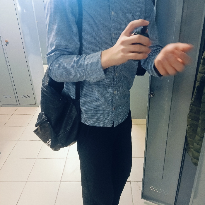 The image depicts a partial view of an individual in what appears to be a locker room. The location is an unspecified facility within the city of Chaikovsky, Russia, as no specific identifying features for an exact address are present.
The subject, an individual of indeterminate gender, is visible from the upper torso to the mid-thigh, oriented slightly towards the right. They are wearing a light blue, small-checkered long-sleeved button-up shirt and dark trousers. A dark-colored backpack or shoulder bag is slung over their left shoulder. The individual's right hand holds a small, dark spray bottle, which is being directed towards their exposed left wrist or forearm. The left hand exhibits slight motion blur, consistent with the act of receiving the spray or possibly rubbing the sprayed area.
The background consists of a row of light-gray metal lockers. At least two lockers on the left side have visible circular, gold-toned handles or locks and small ventilation grilles near their bases. On the far right, one locker door is partially open, revealing a dark green, possibly quilted, garment hanging inside. The floor is tiled with light-colored square tiles.
