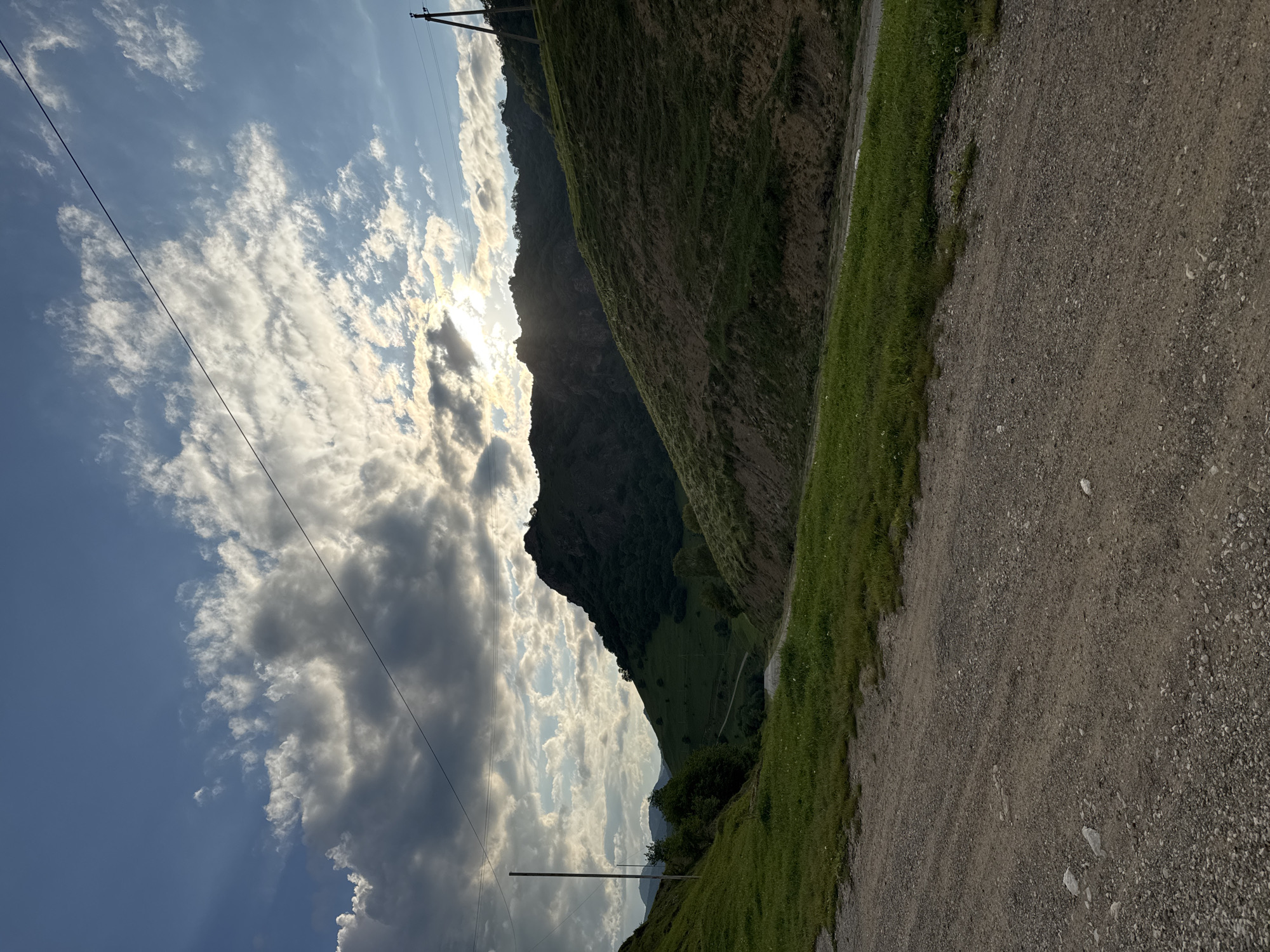 This media file depicts a scenic view of a mountain landscape with a dirt road in the foreground. The scene is dominated by the rugged slopes of a mountain covered in green grass and sparse trees. A winding dirt road, bordered by a strip of vibrant green grass, runs along the right side of the frame. In the background, towering mountains rise, their dark, rocky surfaces contrasting with the bright, cloudy sky.
The sky is a mix of blue and white, with dramatic, sun-drenched clouds creating a sense of depth and light. The sun is partially obscured by the clouds, suggesting either early morning or late afternoon light. The overall mood is serene and natural, highlighting the raw beauty of the mountainous terrain. There are visible power lines stretching across the sky, indicating the presence of some infrastructure, but they do not detract from the natural splendor.
The location is identified as Nizhnyaya Mara, Russia, which provides context for the mountainous and possibly remote setting. The image captures a moment of natural tranquility, with no people or specific activities taking place, allowing the viewer to appreciate the expansive landscape and atmospheric conditions. The textures of the dirt road, the grassy slopes, and the cloudy sky are all rendered with detail, contributing to the immersive quality of the image.
