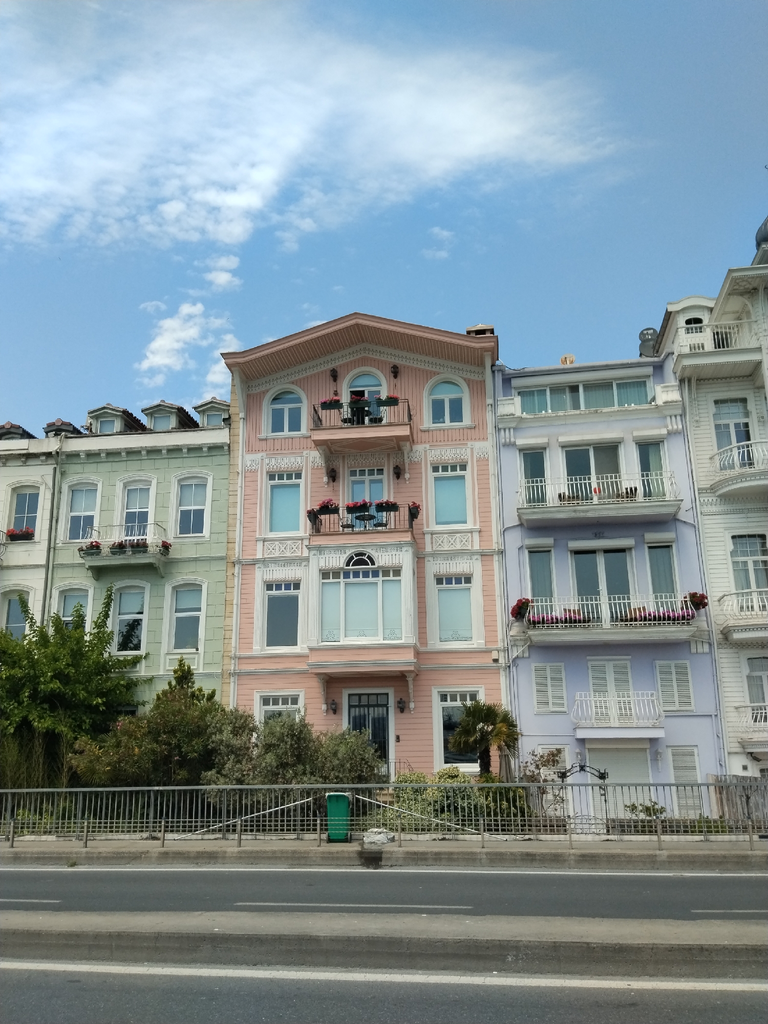 The image displays a picturesque street scene featuring a row of ornate multi-story residential buildings in Beşiktaş, Turkey, under a clear blue sky with scattered white clouds. The buildings are painted in distinct pastel colors: light green on the left, a prominent pink building in the center, and a light purple building, with a portion of a white building visible on the far right.
Each building exhibits intricate architectural details, including decorative cornices, window frames, and balconies with white railings. Many balconies are adorned with potted red and pink flowers, adding vibrant accents to the facades. The central pink building features a distinctive arched pediment and bay windows. Dormer windows are visible on the roofs of the leftmost buildings, and satellite dishes can be seen on some rooftops.
In front of the buildings, a lush line of green trees and bushes separates them from the foreground. A sturdy metal fence runs across the mid-ground, acting as a barrier between the greenery and the road. A green trash can is visible behind the fence on the right side. The immediate foreground features a paved asphalt road with a concrete median and white road markings.
The scene appears to be set during the daytime, likely under pleasant weather conditions. No people or specific activities are visible, presenting a tranquil urban landscape. There is no visible text in the image.