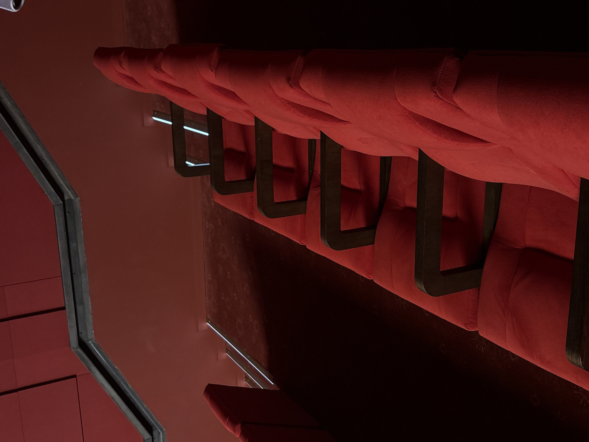 The image depicts a row of unoccupied red upholstered seating in what appears to be a theater or auditorium. The seating has dark, possibly wooden armrests and the upholstery is textured. The environment surrounding the seating includes red walls and a floor. The perspective and angle of the shot are slightly skewed. This is possibly a location in the city of Minsk, Belarus.