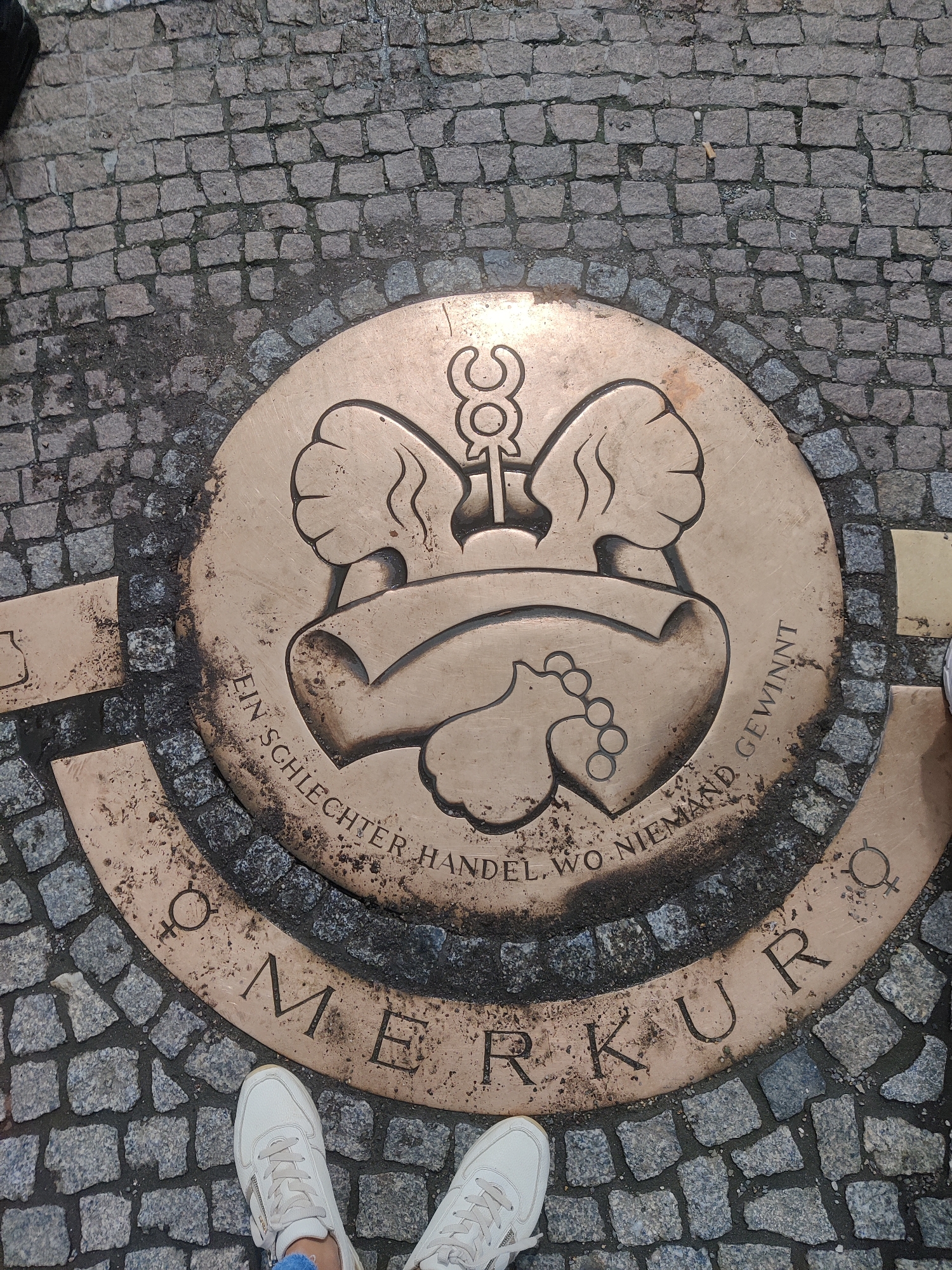 A close-up, eye-level shot shows a circular bronze plaque embedded in a cobblestone street. The plaque features a relief of two hands clasped together, forming a heart shape, with a caduceus symbol above. The text around the plaque reads "EIN SCHLECHTER HANDEL, WO NIEMAND GEWINNT" and "OMERKUR". This inscription and imagery refer to the "Market Mercury" (Marktmerkur), a monument dedicated to the spirit of fair trade, located at the Neumarkt in Dresden, Germany. Two pairs of white sneakers are visible at the bottom of the frame, suggesting the viewpoint of a person standing on the street. The cobblestones are grey and show signs of wear and tear, indicating an outdoor, public space.