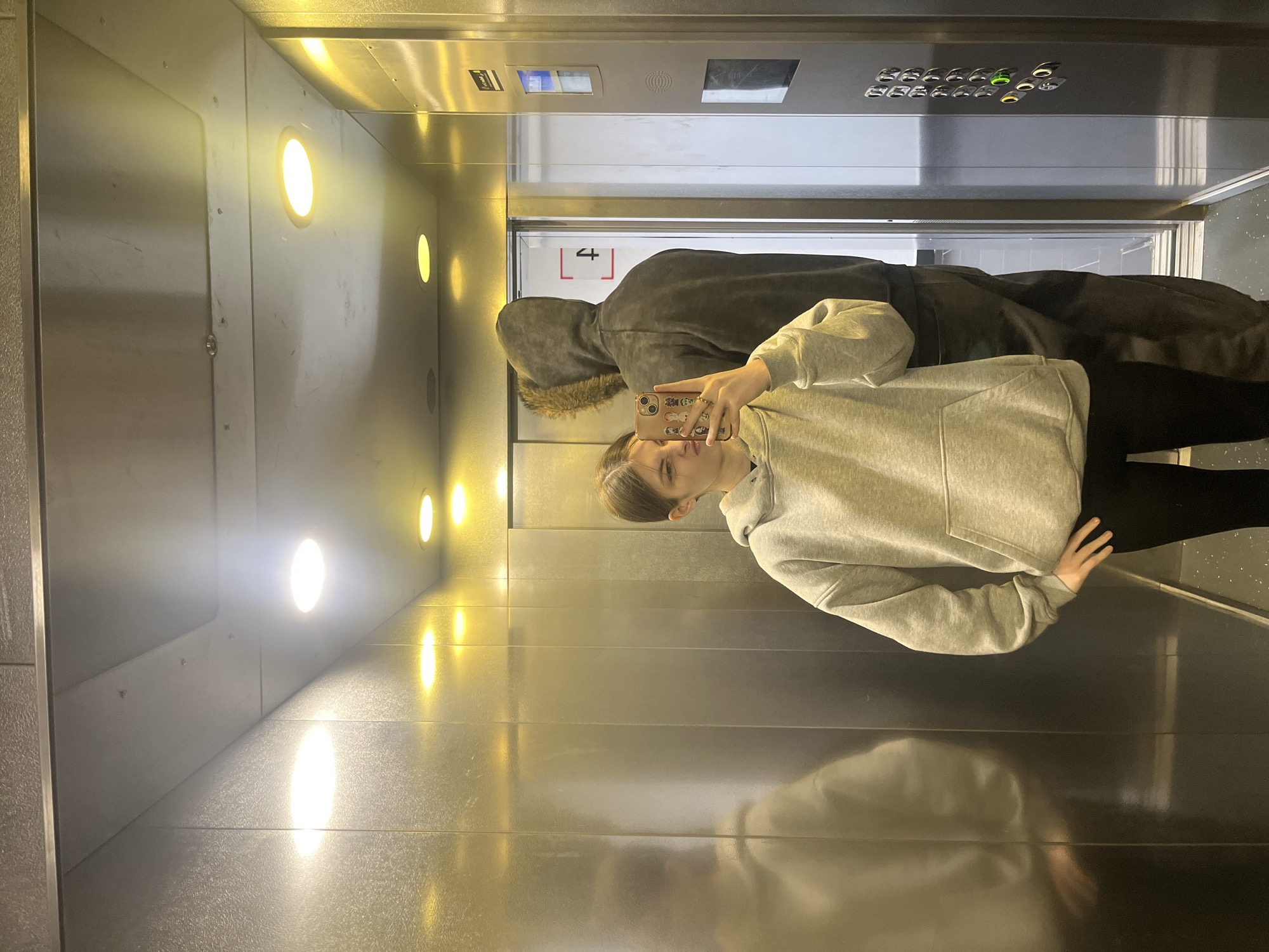 A young woman is taking a selfie in an elevator with shiny metal walls. The elevator has recessed circular lights on the walls, and a panel of buttons above. A person in a dark hooded jacket stands next to her. The location is Saint Petersburg, Russia based on a sign with Cyrillic characters visible in the background.