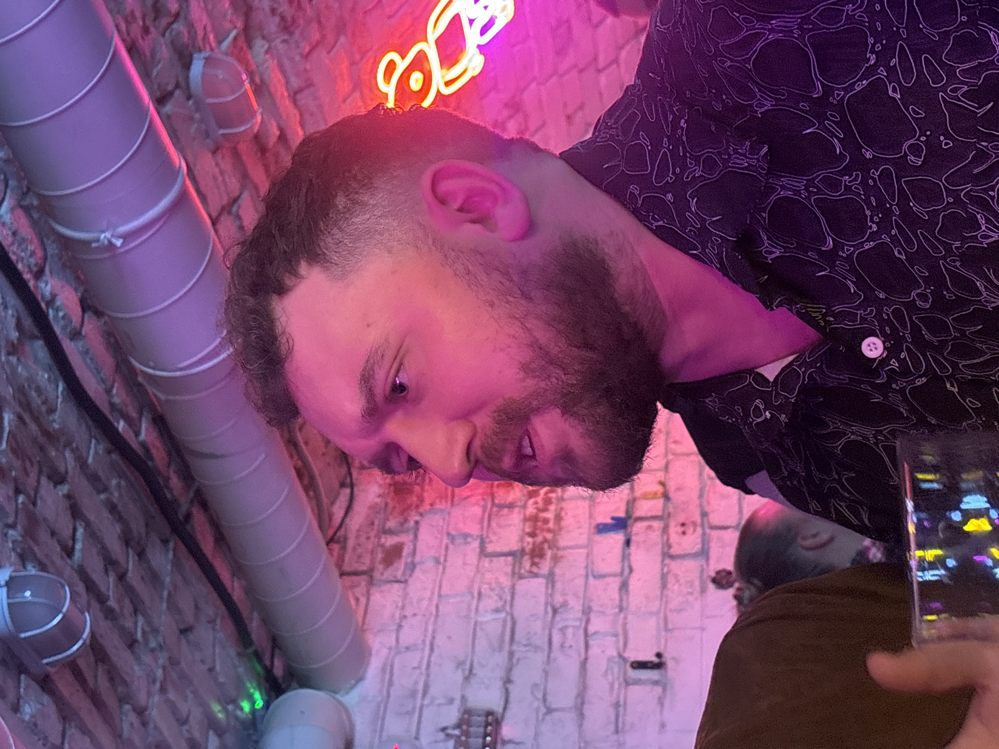 The image shows a man with a beard, wearing a black patterned shirt, in an indoor location. The background features a white-painted brick wall with a neon sign. A large, cylindrical ventilation duct is running along the wall. Another person's head is partially visible, and an item resembling a phone or glass is in the lower right corner. The lighting appears to be predominantly pink and purple. The city of Krasnoyarsk, Russia is not confirmed or evident from the information provided in the image.