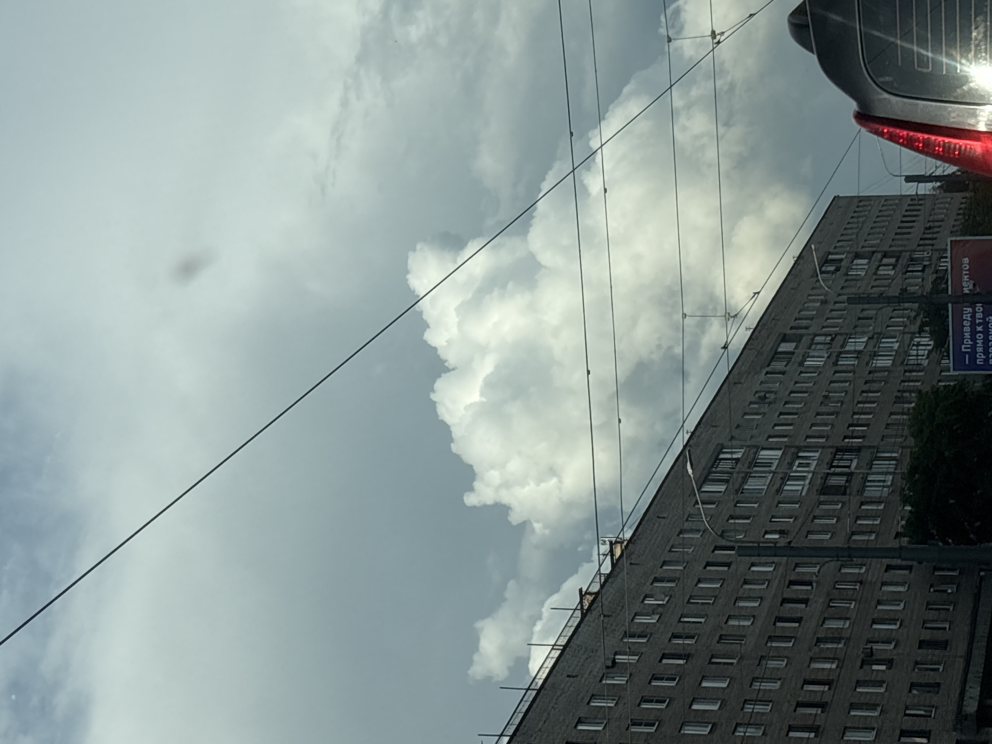 The image captures a view of a large, multi-story brick apartment building in Saint Petersburg, Russia, under a dramatic, cloudy sky. The building appears to be an older Soviet-era structure, characterized by its repetitive window pattern and utilitarian design. Several overhead wires, likely for trolleybuses or trams, crisscross the frame, obscuring parts of the sky and the building. In the foreground, the rear of a dark-colored vehicle is visible, with its red taillight illuminated. The sky itself is a mix of dark, heavy clouds and brighter, cumulus formations, suggesting a dynamic weather system. A billboard is partially visible in the lower right corner, with Russian text. The overall scene conveys a sense of urban life with a focus on the architecture and the prevailing weather conditions.