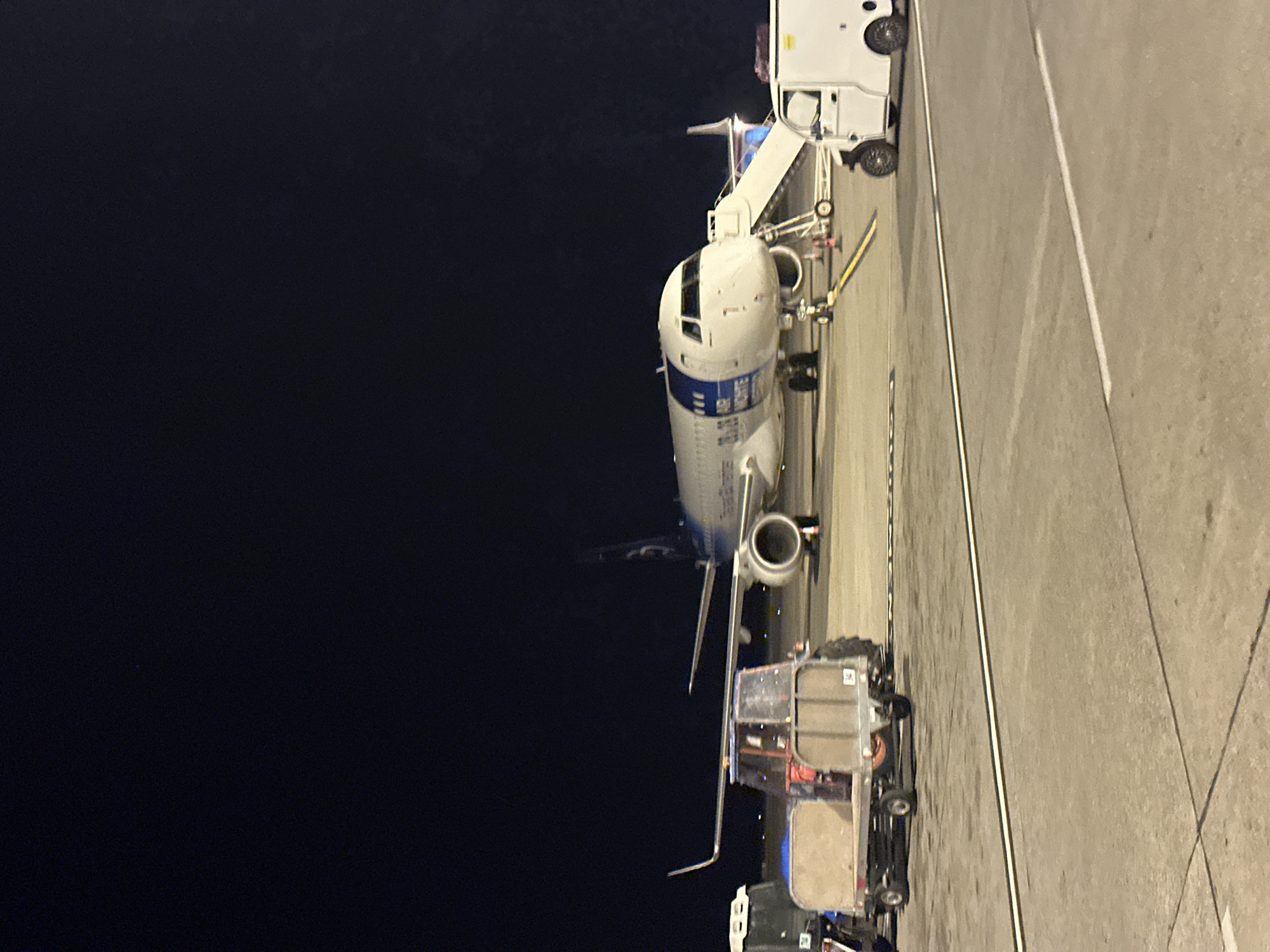 The image depicts an airplane at night on a tarmac, likely at Podgorica Airport in Montenegro, though this cannot be confirmed. The plane is connected to a jet bridge. A baggage handling vehicle is also present, parked near the aircraft. The overall environment suggests an airport setting. The lighting is artificial, indicating nighttime. The scene appears static, without any visible people or active operations. The image is taken at an angle.