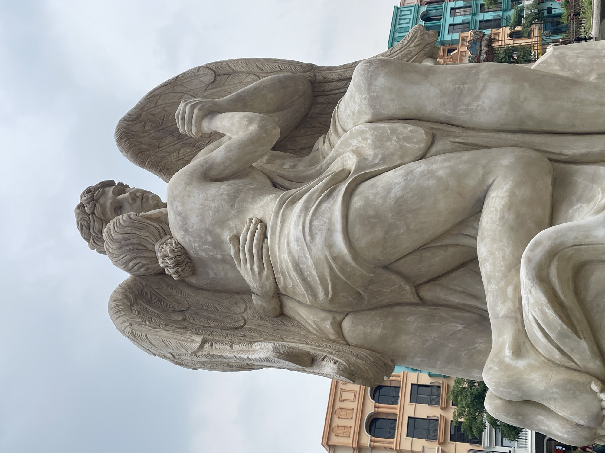 This is a medium shot, eye-level, outdoor photograph of a large, light-colored stone statue of two winged figures embracing. The statue depicts two figures with wings, one kneeling and embracing the other, who is seated. Both figures are partially draped in flowing fabric. The statue is positioned in front of a backdrop of colorful, European-style buildings and a cloudy sky. This statue is located at the Sun World complex in Nghĩa Trụ Commune, Vietnam.