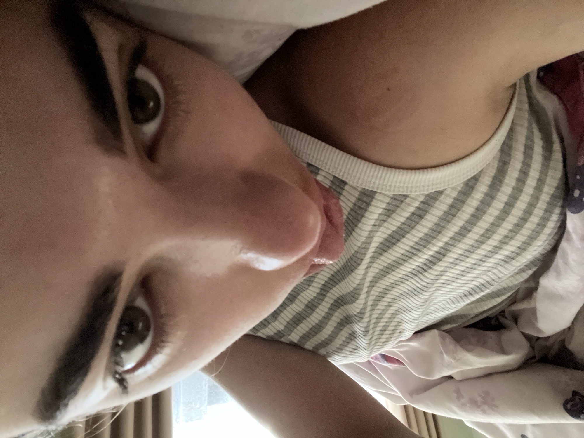 A close-up, low-angle shot shows a person lying on a bed. The person is wearing a striped t-shirt. Their face is the primary focus, with one eye and part of their nose and mouth visible. The background includes bedding and what appears to be a window with vertical blinds. The scene is indoors. There is no indication of the location in Ust-Chorna, Ukraine.