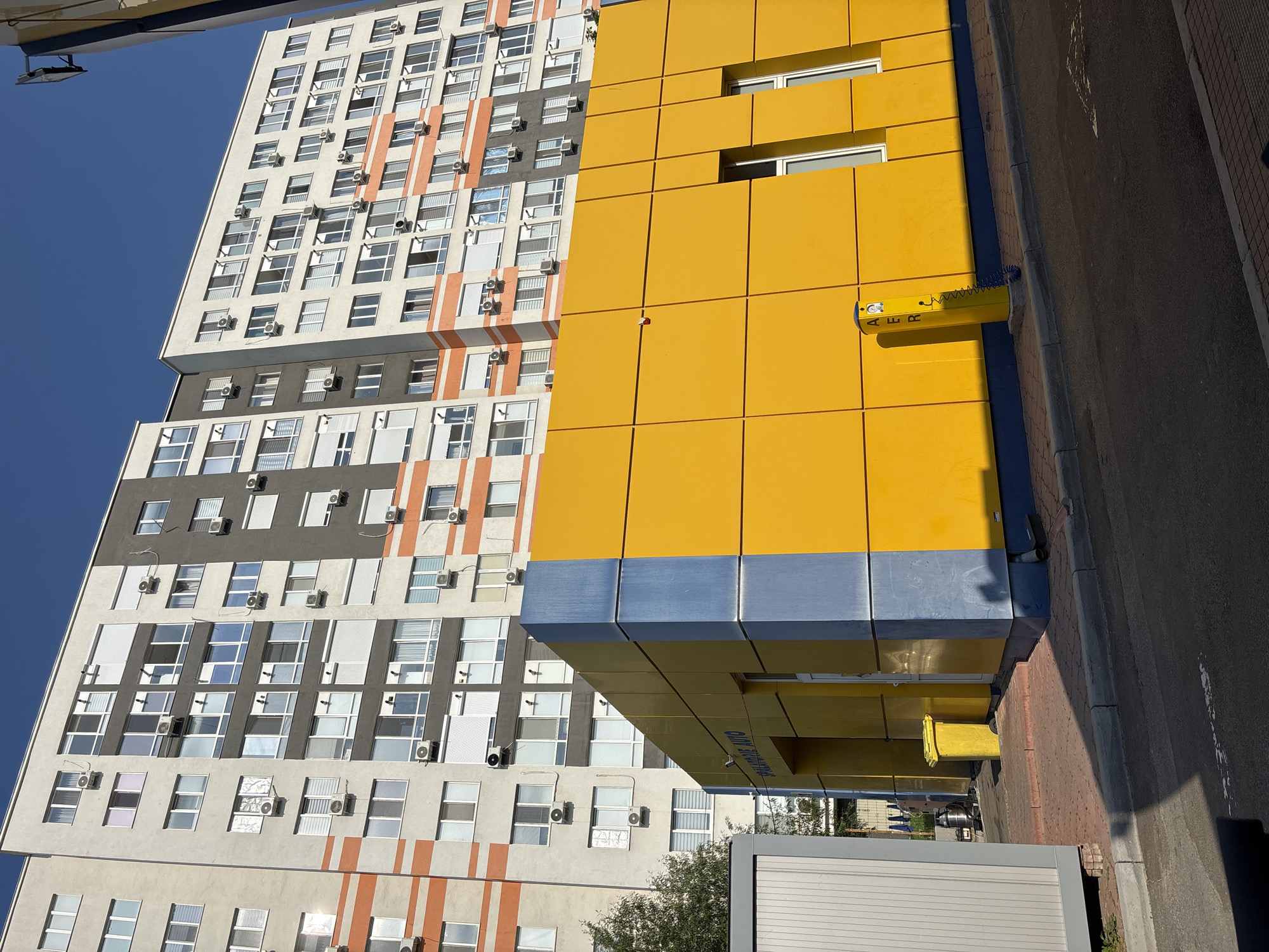 An exterior, eye-level, medium shot captures a modern, multi-story building and a bright yellow structure in an urban environment under a clear blue sky. The multi-story building, clad in light gray with horizontal orange and dark gray accents, features numerous windows and air conditioning units. In the foreground, a contemporary, low-rise building with a striking yellow facade and a blue vertical accent element is visible. A yellow trash bin sits near the entrance of the yellow structure. To the left, a portion of a gray modular structure is present. The ground is paved with red brick and asphalt. A yellow pneumatic stand is positioned along the sidewalk. The scene suggests a commercial or residential area with modern architecture.