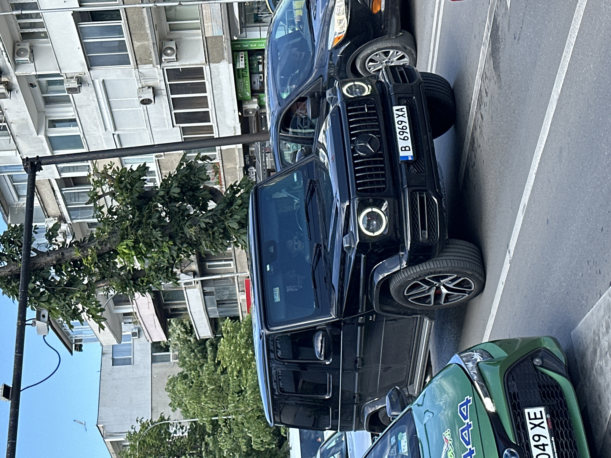The image shows a street scene in Varna, Bulgaria. A black Mercedes-Benz G-Class with license plate "B 6969 XA" is parked in a parking space along the street. The car is positioned between a dark-colored SUV and a green Toyota Corolla with license plate "B 049 XE". The street is lined with buildings, one appearing to have multiple residential units with air conditioning units attached to the exterior. Trees are visible on the street, partially obscuring the view of the buildings. A black utility pole stands on the left side of the frame. The sky is visible and appears clear.