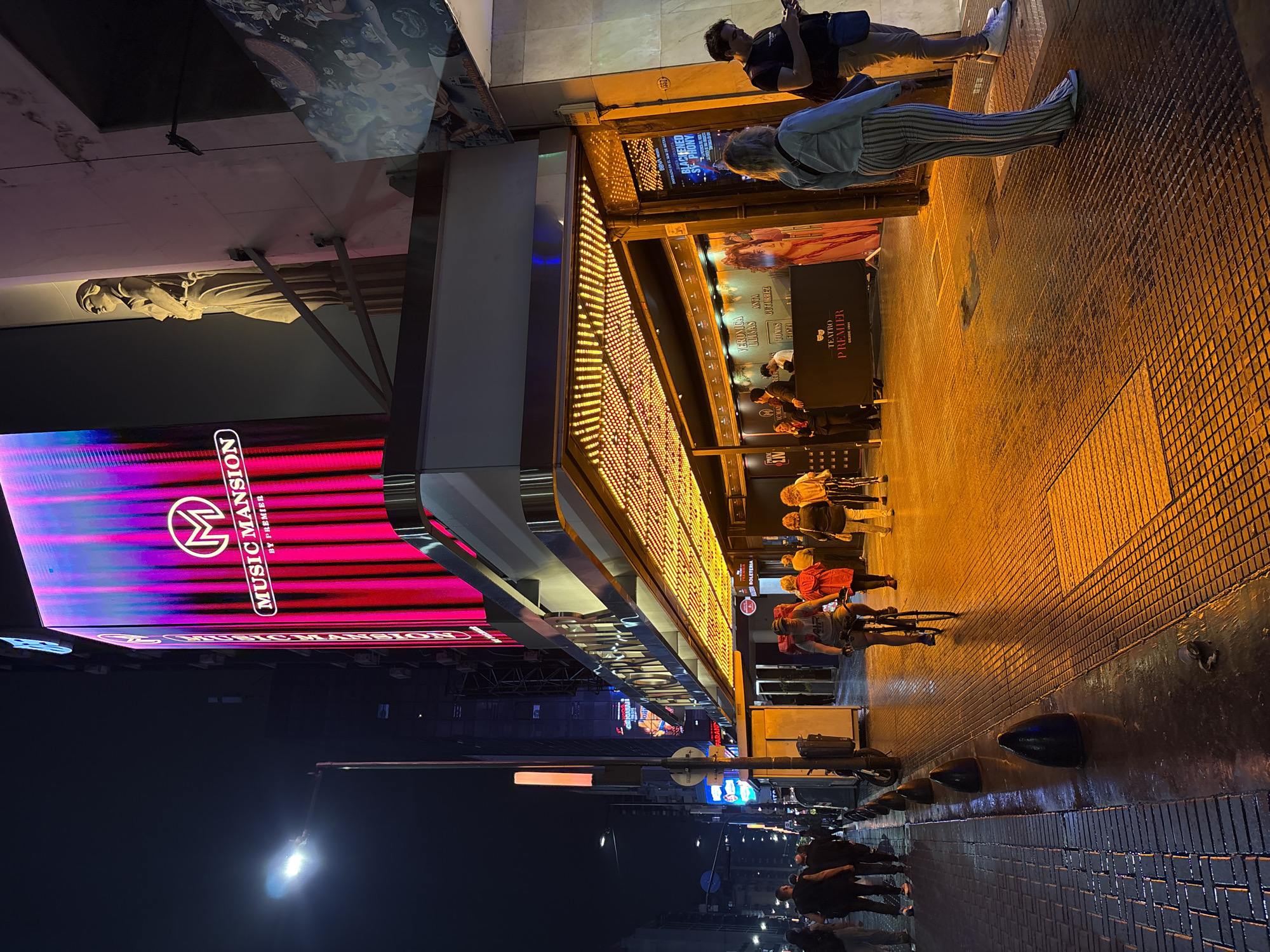 The image depicts a nighttime street scene on Corrientes Avenue in Buenos Aires, Argentina, centered on the facade of the Teatro Premier. The multi-story building features a large, vertical LED screen displaying "MUSIC MANSION by PREMIER" with a circular "M" logo against a backdrop of animated vertical pink, purple, and red light stripes. To the right of the screen, a classical-style female statue is mounted high on the building's light-colored wall. Below the LED screen, an ornate marquee extends over the sidewalk, illuminated by numerous small yellow lights. The area directly beneath the marquee, including the sidewalk, is bathed in a warm, orange light.
Several individuals are present on the sidewalk. Approximately 15-20 people are visible. In the mid-ground, a person wearing a cap and t-shirt rides a black bicycle towards the viewer, positioned on the textured metal grate section of the sidewalk. Near the theater entrance, a group of people stands; one individual in a dark uniform is visible near a "TEATRO PREMIER" branded barrier. Show posters and signs are visible, including "VERONICA LUKAS" and "BLACKENED SYMPHONY," along with "BOLETERIA" (ticket office). In the foreground right, a person in a light blue jacket and vertically striped light-colored pants stands with their back partially to the viewer, next to a male in a dark t-shirt and light pants who is looking at a mobile phone.
The sidewalk comprises a mix of grey brick pavers and darker textured metal grates, appearing wet and reflective, suggesting recent rain or dampness. Black, conical bollards line the curb on the left side of the street. A bright street light illuminates the upper left portion of the scene. In the background, other illuminated buildings and streetlights line the avenue. A crushed aluminum can lies on the wet ground near the curb. A light-colored kiosk or electrical box is situated on the left sidewalk, behind the bollards.