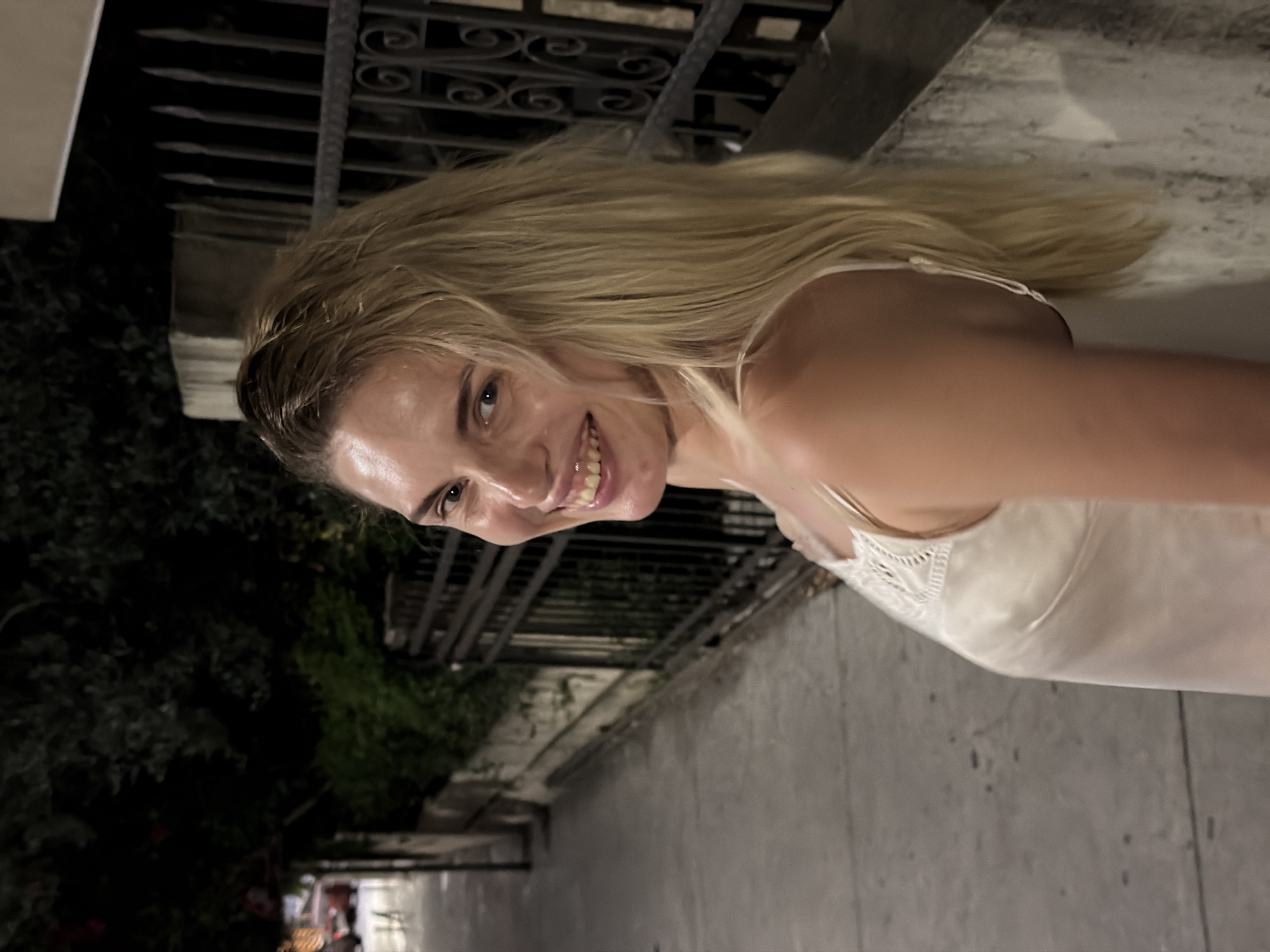 A woman with blonde hair is looking over her shoulder and smiling at the camera. She is wearing a white slip dress with lace detailing on the straps. The background shows a nighttime street scene with a sidewalk, a fence, and some trees. The location is identified as Budva, Montenegro.