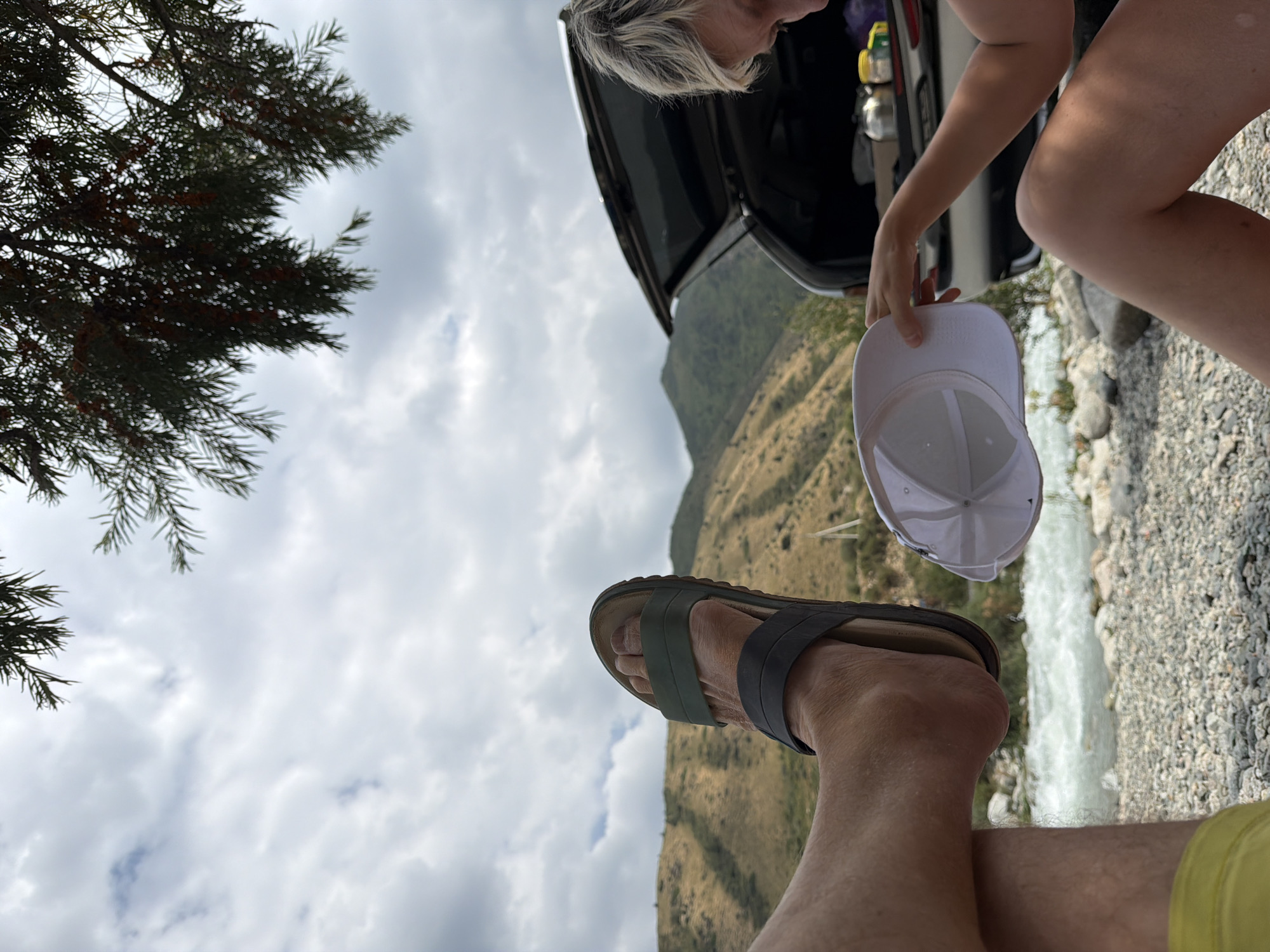 An outdoor scene depicts a person's leg and foot wearing a sandal in the foreground. Behind it, another person is visible, holding a white cap. In the background, a car with an open trunk is parked, and beyond that, a river flows past a rocky shore and hills. The sky is partly cloudy. The location is Unknown, Unknown.