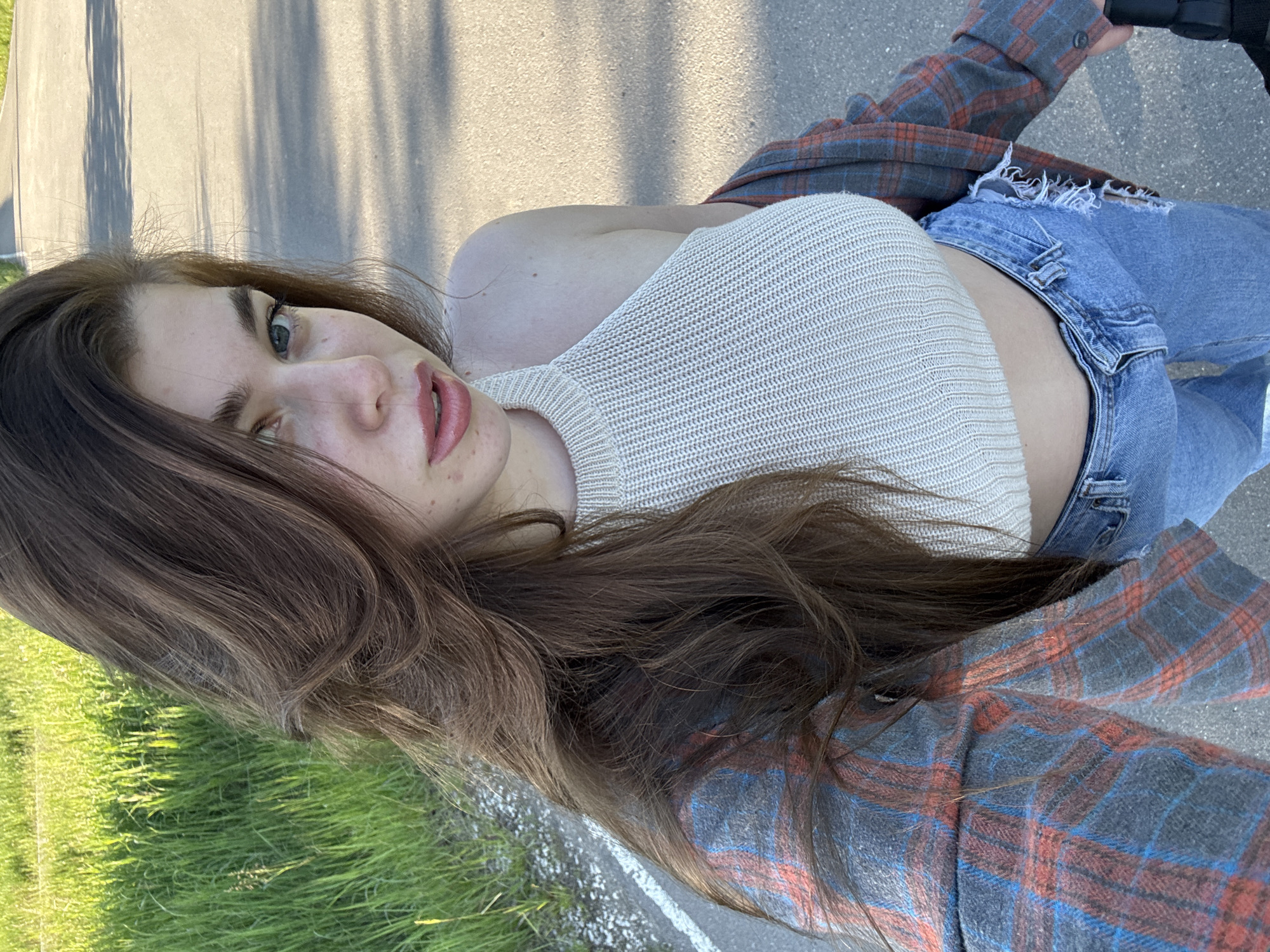 A young woman with long, wavy brown hair, blue-green eyes, and light pink lips is featured in a self-portrait, captured from a slightly elevated and tilted perspective. She is wearing a light-colored, possibly off-white or beige, knitted halter top that exposes her shoulders, and light blue ripped jeans. A plaid flannel shirt, with a pattern of grey, red, and blue checks, is casually draped over her left arm and shoulder.
The setting is outdoors on what appears to be a sunny day, evidenced by bright natural light and distinct shadows cast on the grey asphalt road in the background. Vibrant green grass is visible on the left side of the frame, suggesting a roadside, path, or park area. A white line marking is also discernible on the pavement. In one of the cropped images, a dark object resembling a bicycle handle is partially visible on the far right, hinting that she may be riding or standing with a bicycle.
Her expression is neutral and candid, looking directly towards the camera. The scene portrays a casual outdoor moment. No visible text is present in the images. The location context indicates this is in Мещёры, Russia.