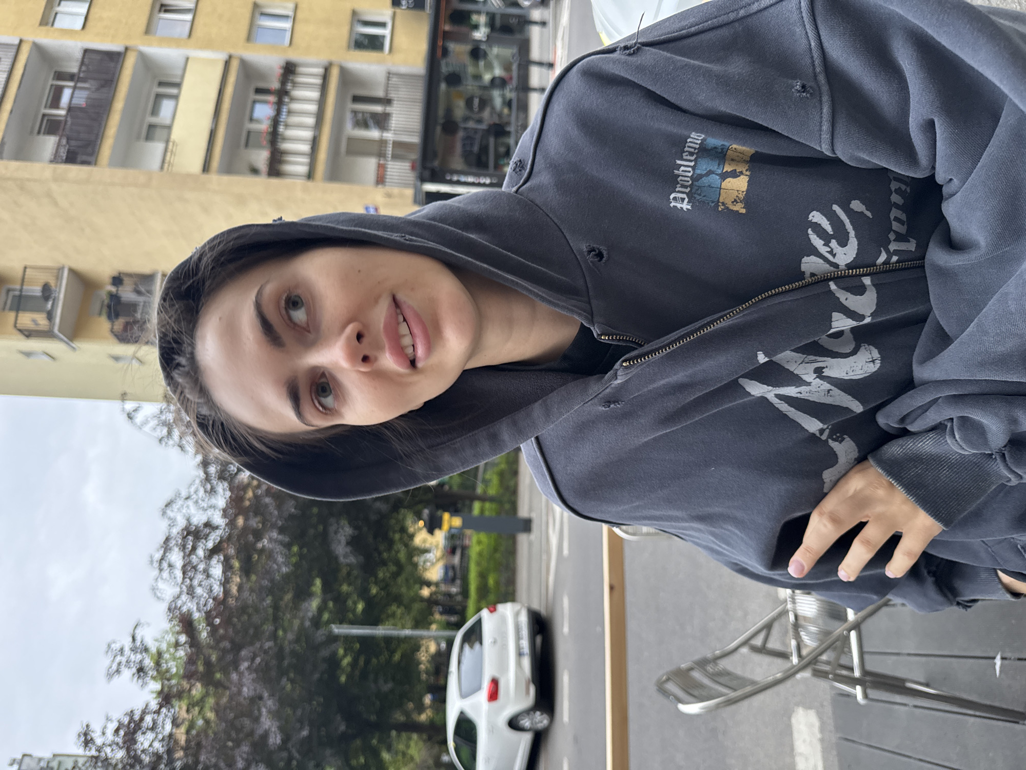 A person wearing a gray hoodie, partially zipped, stands on a city sidewalk. The hoodie has "Problems" written on the left upper chest and a larger design below the zipper. The person looks upward and to the left. The sidewalk is next to a street with a white car. A metal chair is partially visible in the lower right corner. In the background is a multi-story yellow building, and trees with dark foliage. The city is Unknown, Unknown.