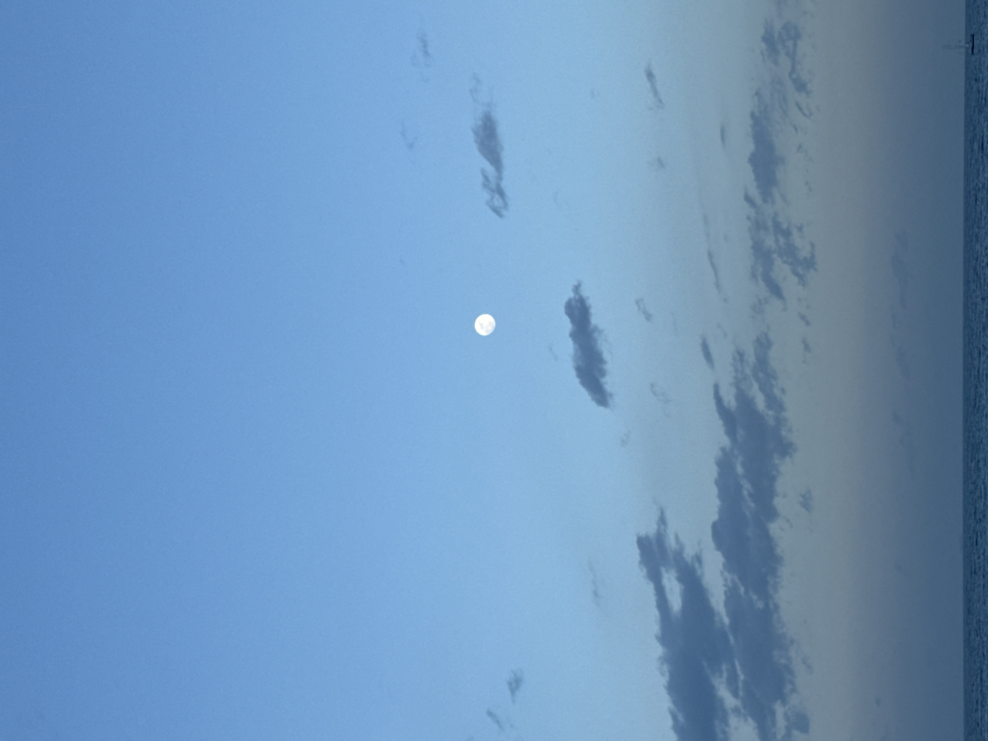 The image presents a daytime scene featuring a bright, full moon visible in the sky. The sky transitions from a light blue hue on the left side of the frame to a slightly faded blue near the right, where clouds are present. The clouds are wispy and elongated. A dark horizontal line along the right edge of the image could indicate the presence of water, potentially the sea. The image is rotated 90 degrees counter-clockwise. The location is not confirmed as being Barcelona.