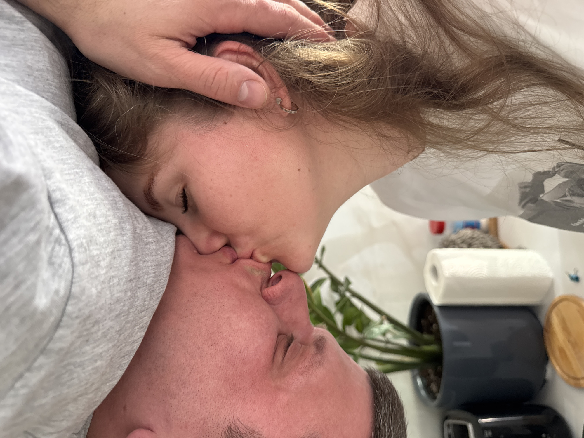 Two people are kissing. A light-skinned woman with brown hair and an earring is kissing a light-skinned man with short, graying hair. The man is wearing a gray shirt. The woman's hand is on the side of her head. In the background is a plant in a dark gray pot, a roll of paper towels, and various other items. The location appears to be indoors. There is no indication that this is in Gornyi, Russia.