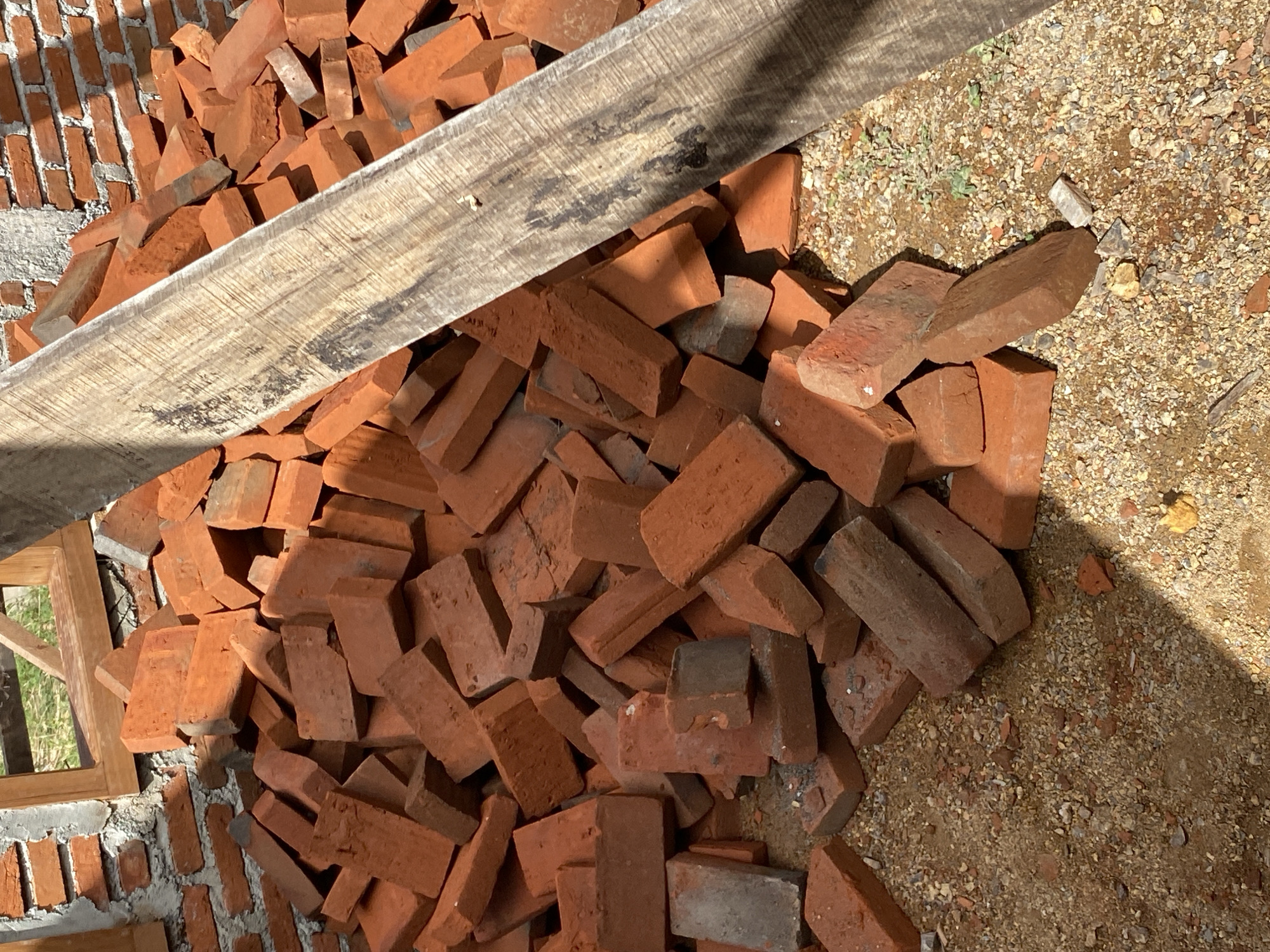 A pile of red bricks is scattered on the ground in front of a partially constructed brick wall. The wall has a wooden frame for a window opening visible. A rough-hewn wooden plank lies diagonally across the foreground, obscuring some of the bricks and casting a shadow. The ground is covered with sand and small pebbles. The scene appears to be a construction site in Banda Aceh, Indonesia.
