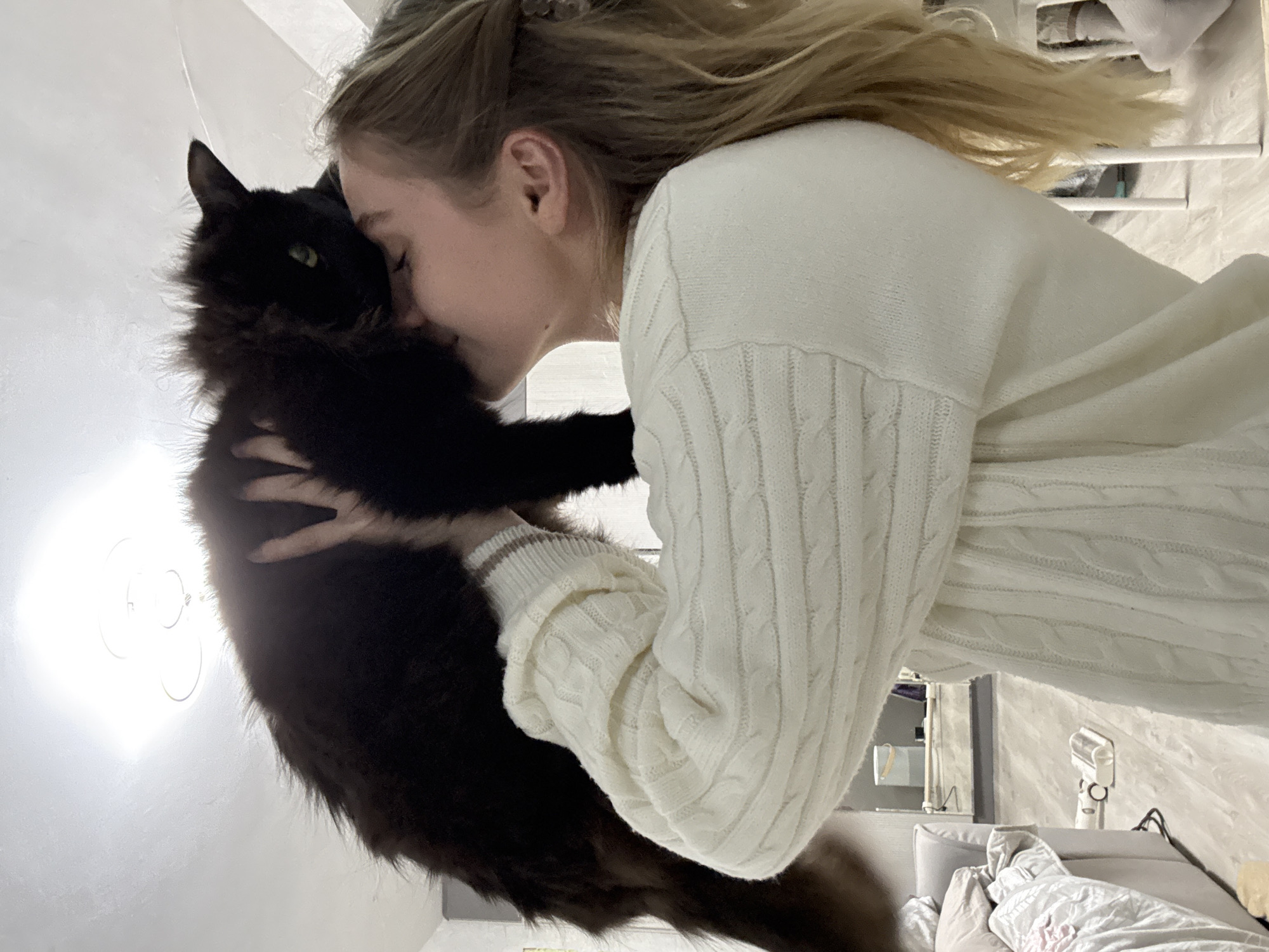 When viewed upright, the image depicts a young woman affectionately embracing a long-haired black cat indoors. The woman, with blonde hair flowing, is wearing a cozy white cable-knit sweater. She holds the cat close to her face, seemingly kissing or nuzzling its head, with her eyes closed, conveying a moment of deep affection and tenderness.
The black cat, with distinct green eyes, is held securely in the woman's arms, its fluffy fur contrasting against her light attire. The interaction highlights a strong emotional bond between the pair.
The setting is a brightly lit indoor space, likely a bedroom or living area. A strong light source, possibly a window or a ceiling light fixture, illuminates the scene from the left. The background features light-colored walls. Towards the right, elements of a room are visible, including a bed with grey pillows and light-colored, patterned bedding. A white vertical appliance, possibly a fan or air purifier, stands on a surface in the background. The overall atmosphere is warm and intimate, capturing a heartfelt connection. The scene takes place during the daytime in Chaikovsky, Russia. There is no visible text.