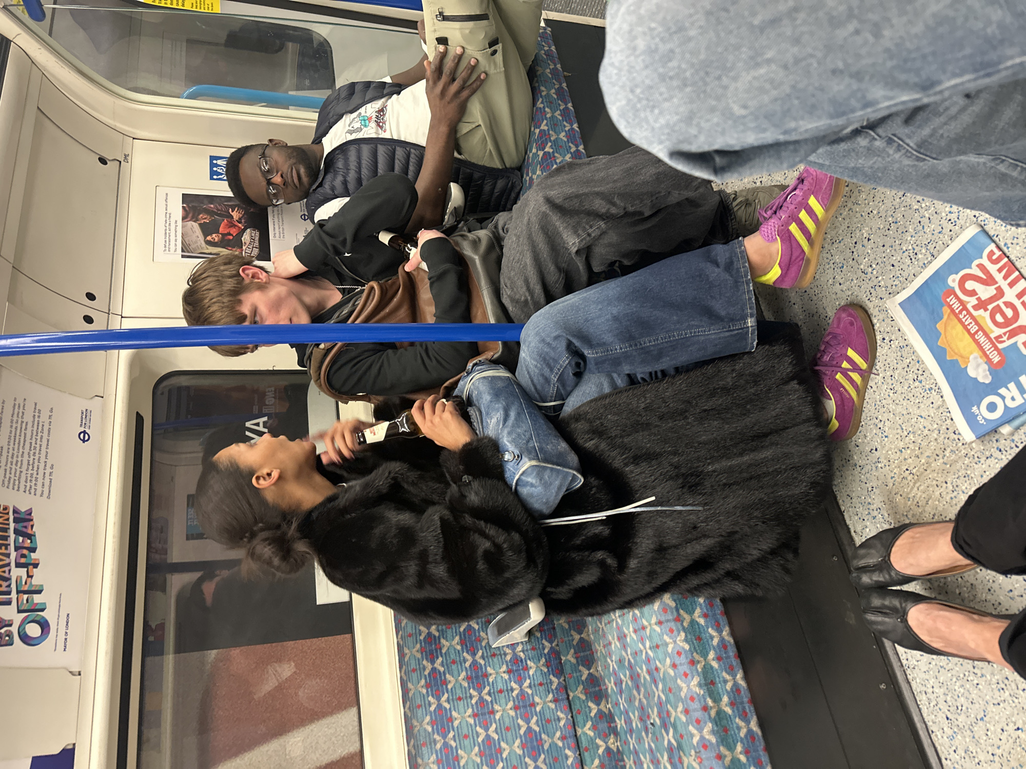 The scene is set inside a London Underground train carriage, indicated by "TRANSPORT FOR LONDON" signage and typical seating patterns. Three individuals are seated on a blue, red, and white patterned bench. On the left, a dark-skinned female with dark hair in a bun wears a black faux fur coat, blue jeans, and purple sneakers with yellow stripes; she holds a brown glass bottle and is speaking, facing the person next to her. A light-skinned male in the center, wearing a black top, grey trousers, and a brown leather shoulder bag, also holds a brown glass bottle and gestures with his left hand near his ear, looking towards the female. To his right, a dark-skinned male with glasses, dressed in a white t-shirt, a dark blue padded gilet, and olive-green cargo trousers, stares directly at the viewer, with his right hand resting on his left thigh. In the lower foreground, the feet of a fourth person are visible, wearing black tabi-style flats and black trousers, positioned on the speckled floor. A newspaper or leaflet displaying "Jet2.co.uk" lies on the floor. Advertisements for "BY TRAVELLING OFF-PEAK" and "MAYOR OF LONDON" are affixed to the white carriage walls.