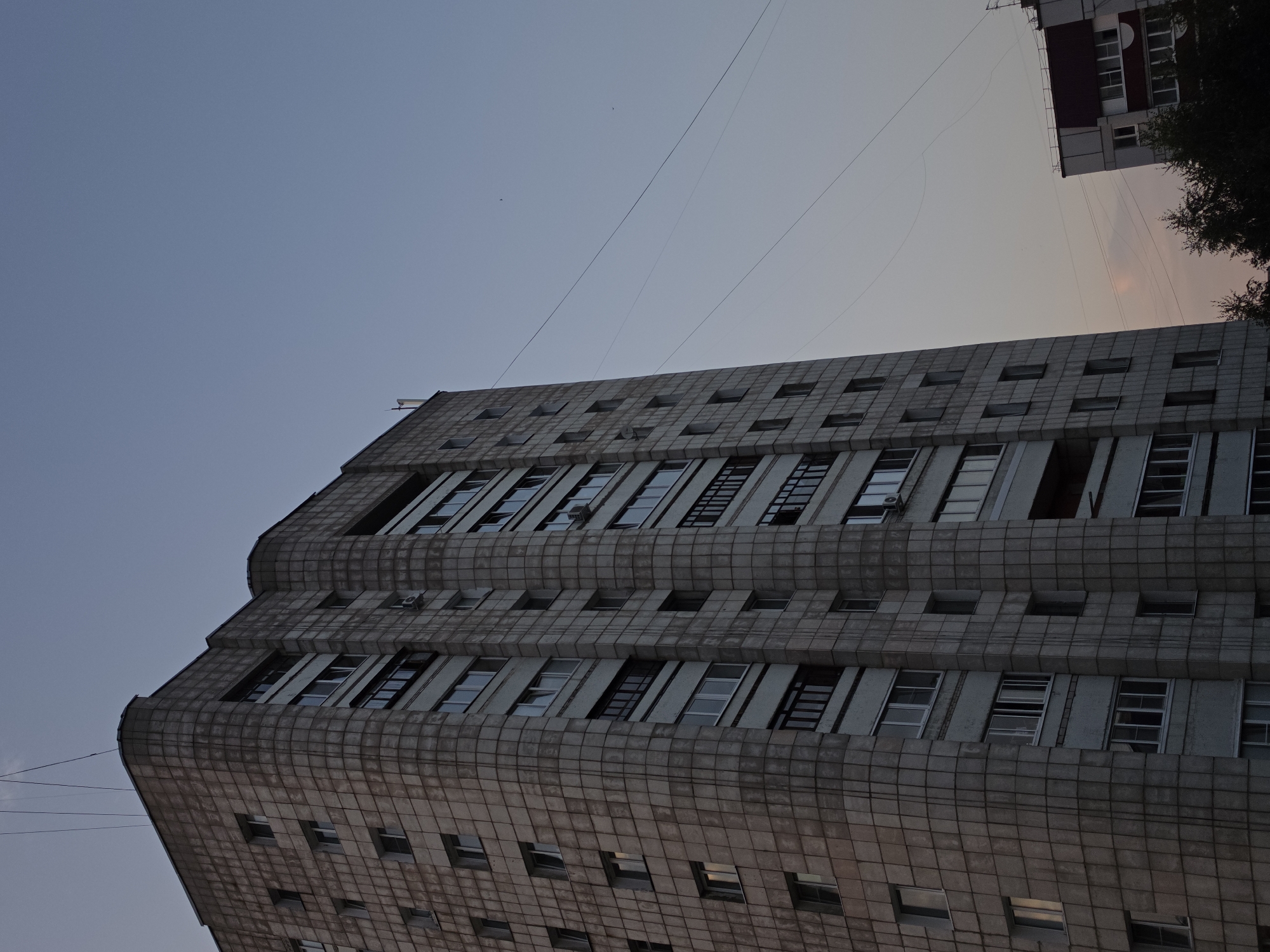 This media file depicts the exterior of a tall, multi-story building. The building's facade is constructed from a grid of square tiles, giving it a textured appearance. Numerous windows are visible, many of which are rectangular and appear to be closed. Some windows have open sections, suggesting ventilation. The building has a curved profile on its visible side.
The scene is set against a clear sky, transitioning from a lighter hue near the horizon to a darker blue overhead. Several thin, dark lines, likely power or telephone wires, stretch across the sky and over the building. In the background, another building and a dark mass of trees are partially visible. The lighting suggests it is either early morning or late evening, as there is no harsh sunlight, and the sky has a soft, diffused glow. No people or specific activities are evident.
The location context provided is Chaikovsky, Russia. There is no visible text in the image.