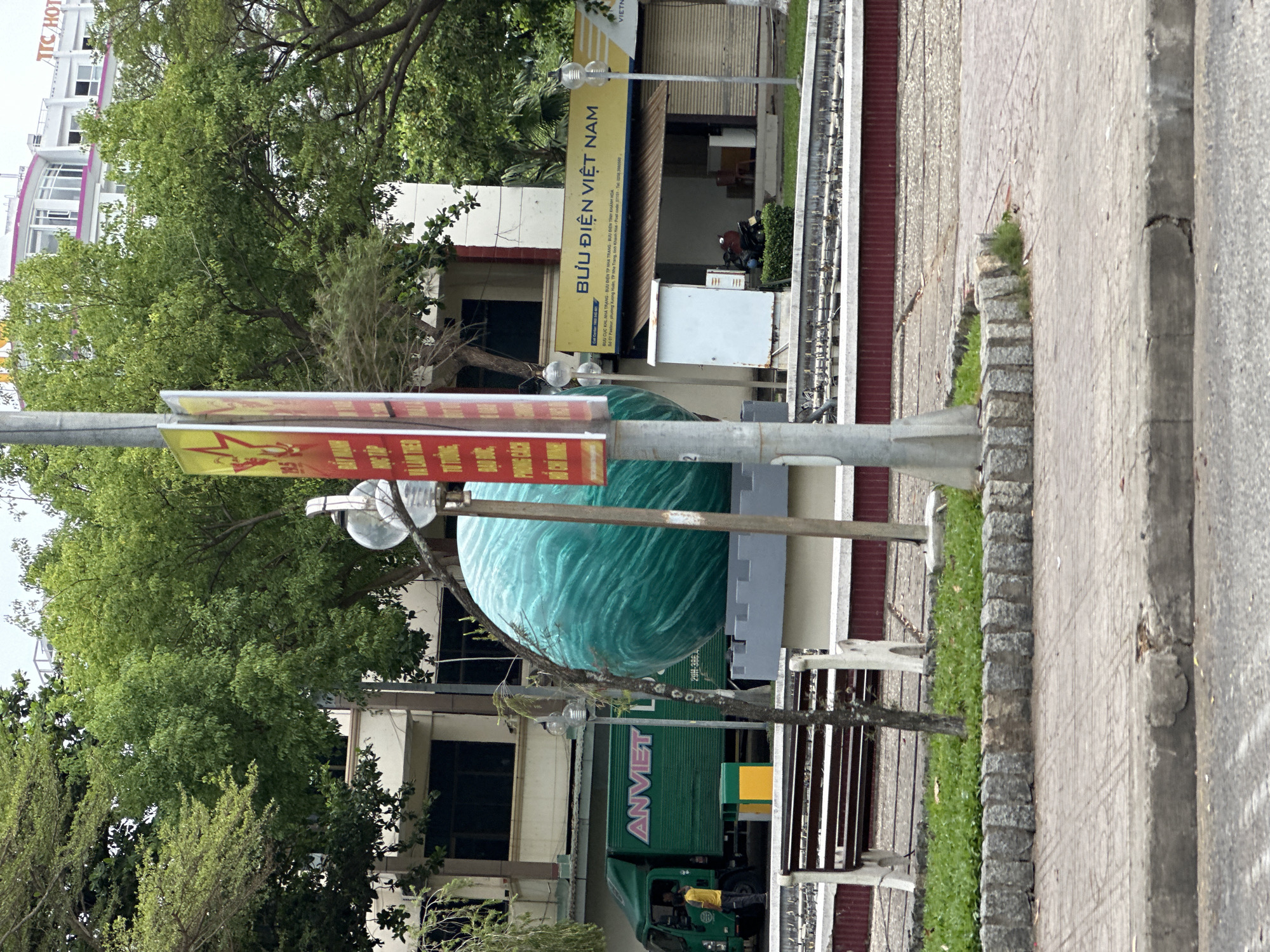 The image shows a street scene in Nha Trang Ward, Vietnam. A truck labeled "ANVIET" is visible on the street. Above the truck is a large, blue, swirling globe sculpture. A sign for "BƯU ĐIỆN VIỆT NAM" (Vietnam Post Office) is visible in the background of the upper right of the frame. Pedestrians are present, including someone near the front of the truck. A sidewalk runs along the right side of the frame. Vegetation and buildings line the street. The scene is outdoors, likely during daylight.