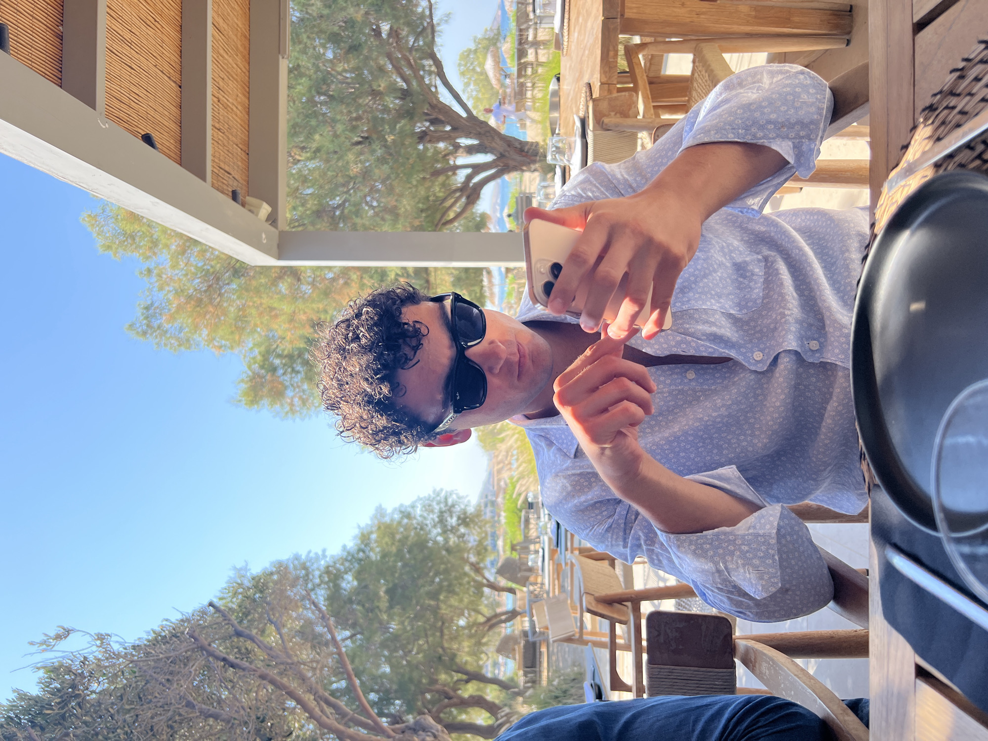 A young man with curly dark hair and sunglasses is seated at a wooden table outdoors, holding and looking at his smartphone. He is wearing a light blue patterned button-down shirt with the sleeves rolled up. The setting appears to be a restaurant or cafe near a beach, with more wooden tables and chairs visible, along with trees and a bright blue sky. In the background, there are glimpses of the sea and beach umbrellas. The overall scene suggests a leisurely day in a Mediterranean location. The specific location in Naousa, Greece is not discernible from the image itself.