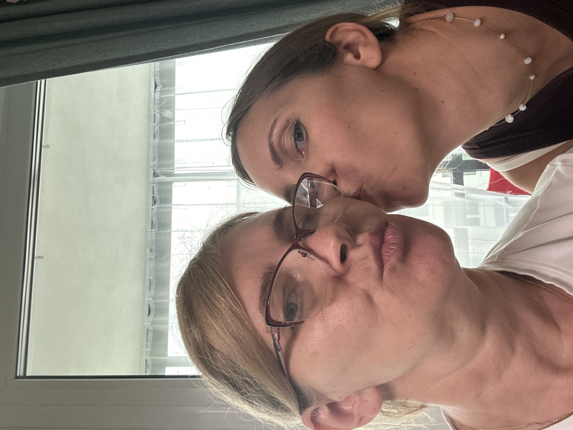 Two women are seen in a close-up, selfie-style shot. The woman on the left is wearing glasses and has her lips pursed. The woman on the right is leaning in to kiss the woman on the left. Both women have light skin and brown hair. They are indoors, in front of a large window with sheer curtains. The background appears to be a domestic setting, possibly an apartment. There is no indication of the specific location in Vsevolozhsk, Russia.