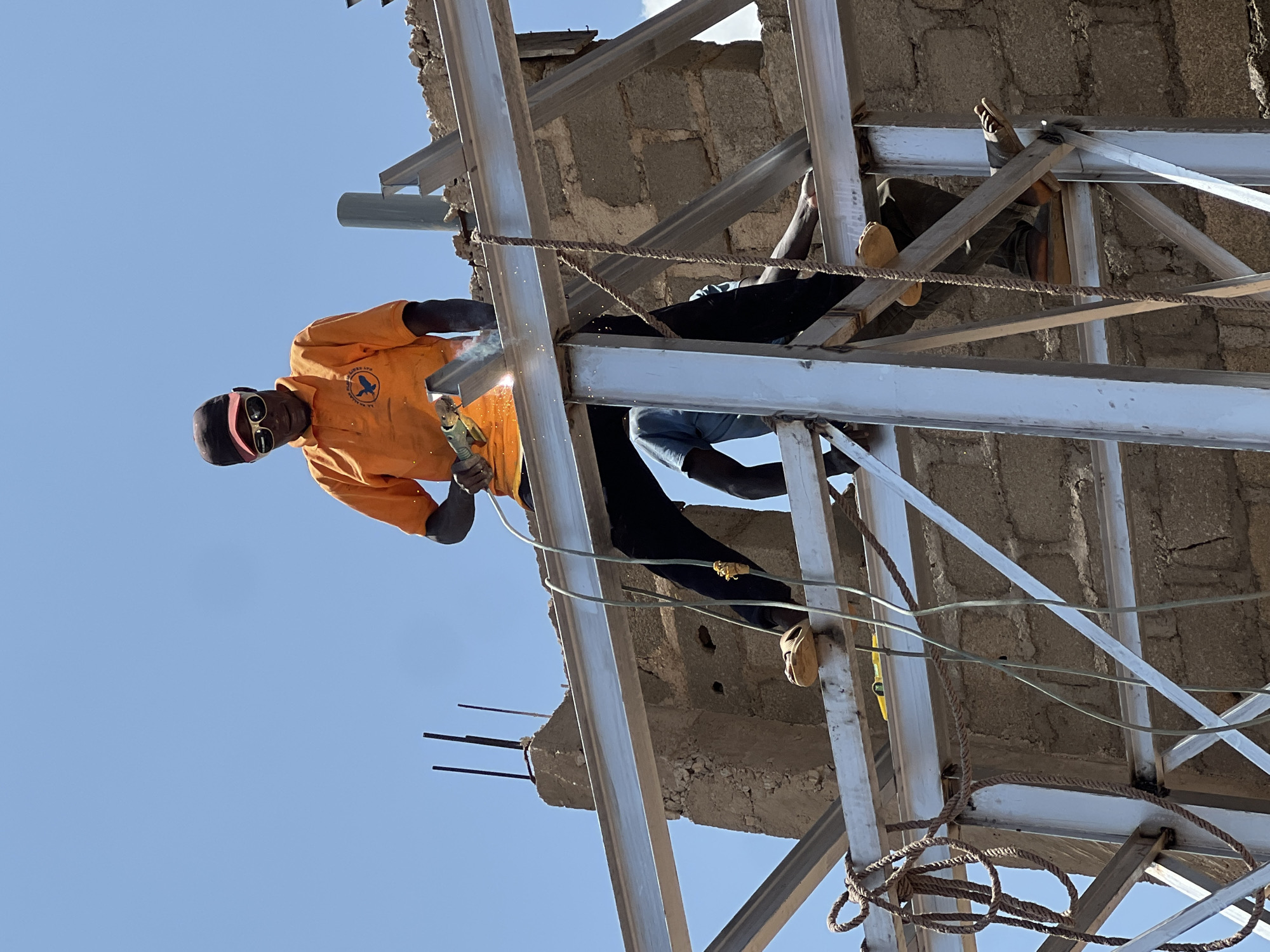 In Jos, Nigeria, under a clear blue sky, construction workers are assembling a metal structure. A man in an orange polo shirt, dark pants, sunglasses, and a red-brimmed cap is actively welding a section of the metal frame. Sparks fly from his welding torch, illuminating his action. Another worker, wearing a blue t-shirt, is visible behind him, assisting with the construction. A third worker, with dark skin and wearing dark pants and sandals, is positioned lower on the structure, also engaged in the building process. Ropes and electrical wires are strung around the construction site, indicating ongoing work. The background reveals a brick wall construction and a grey pipe protruding from a concrete pillar, suggesting the initial stages of building development.